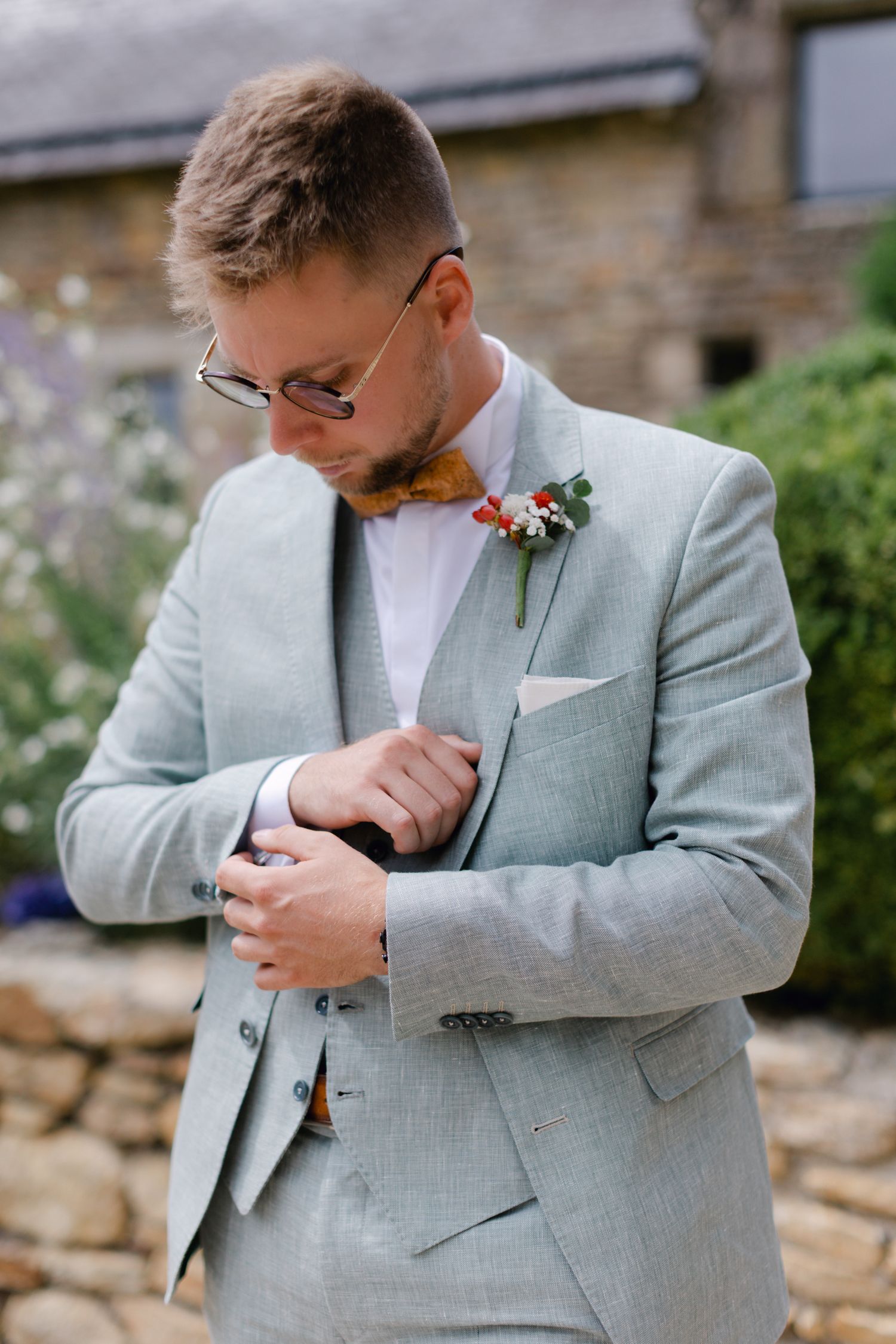 Marié dapper en costume gris clair ajustant les boutons de manchette avec nœud papillon en bois et boutonnière.