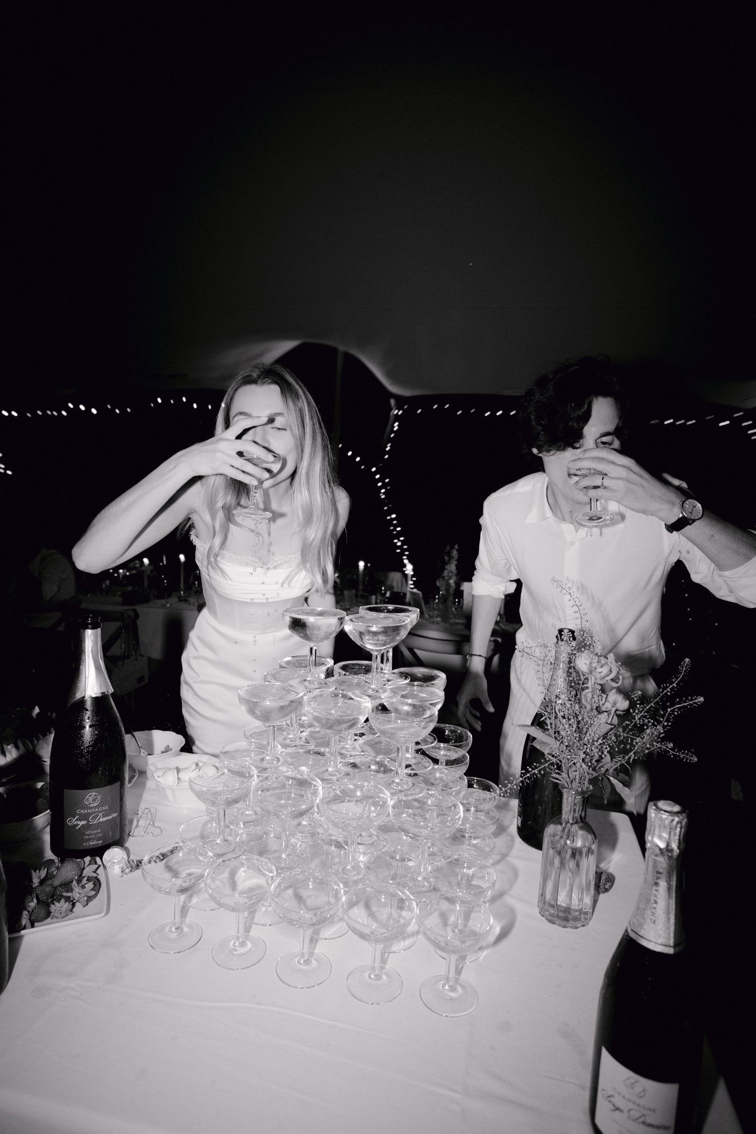 Photo en noir et blanc de la mariée et du marié riant à côté de la tour de champagne au mariage.