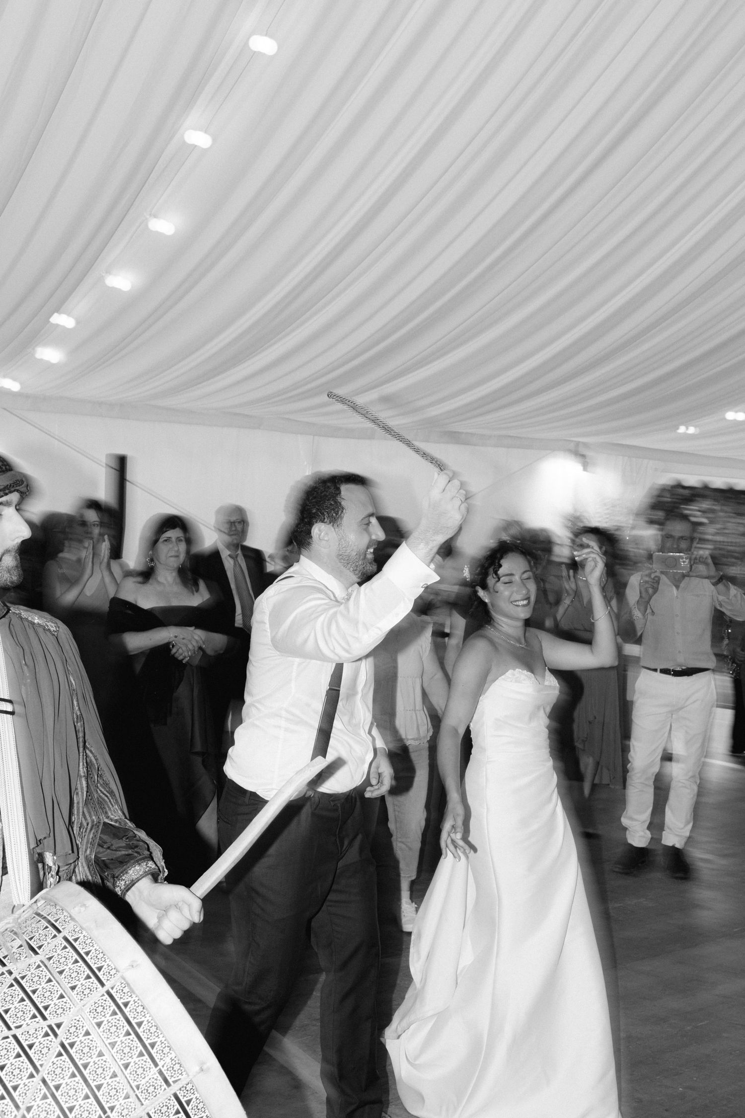 Black and white photo of bride and groom dancing joyfully at wedding reception under draped ceiling.