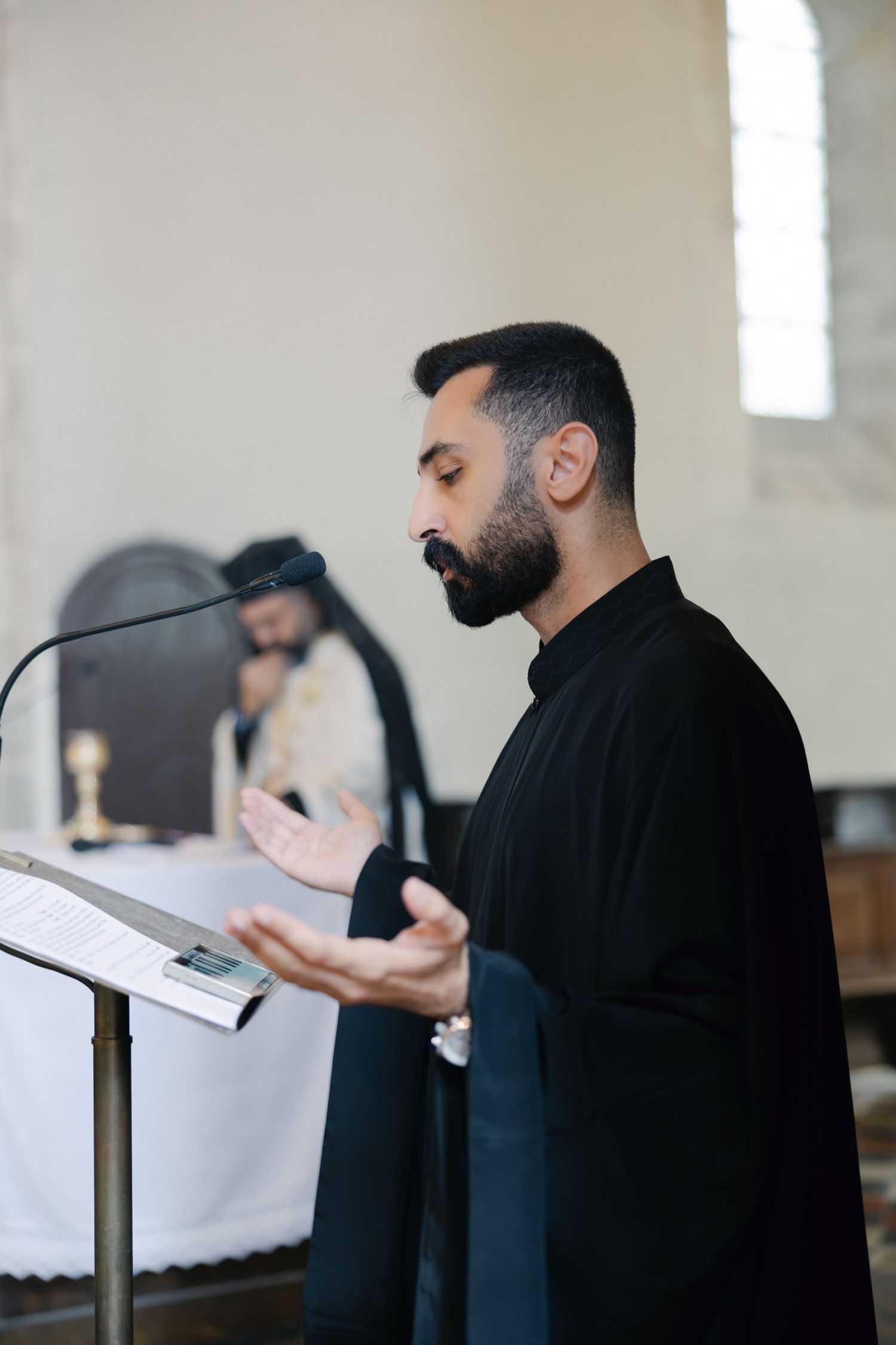 Bearded man in black clergy attire praying at a lectern inside a church during a religious service.