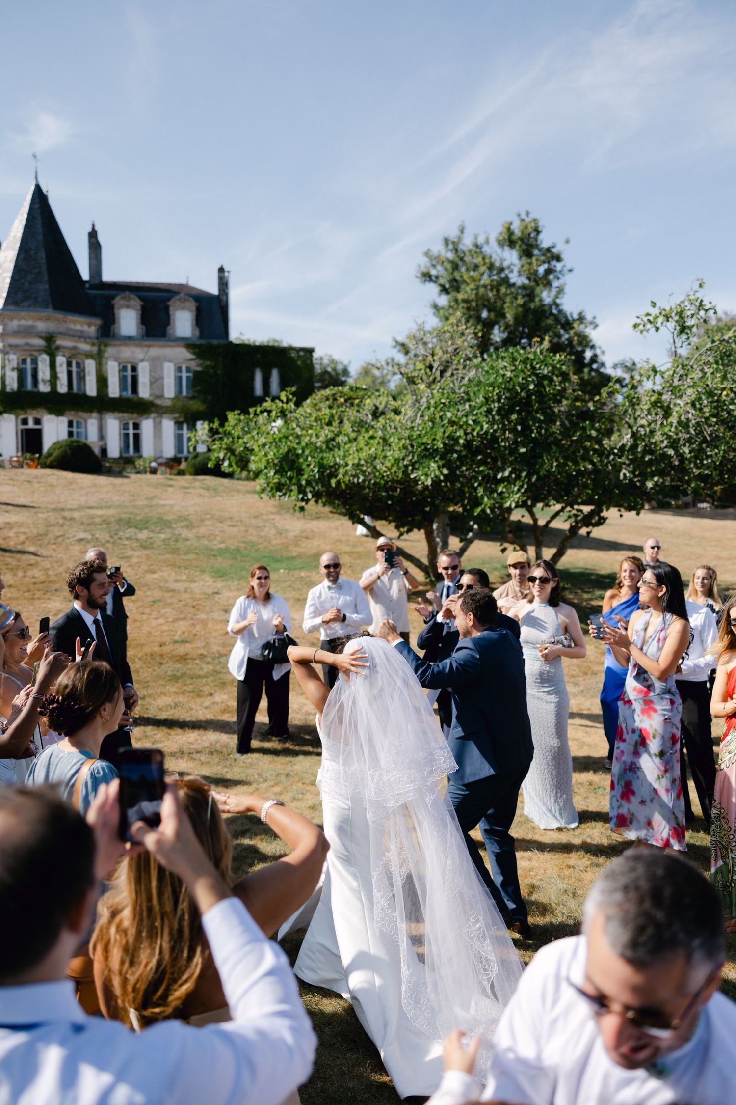 Bride and groom share first kiss outdoors at elegant French chateau wedding surrounded by cheering guests.