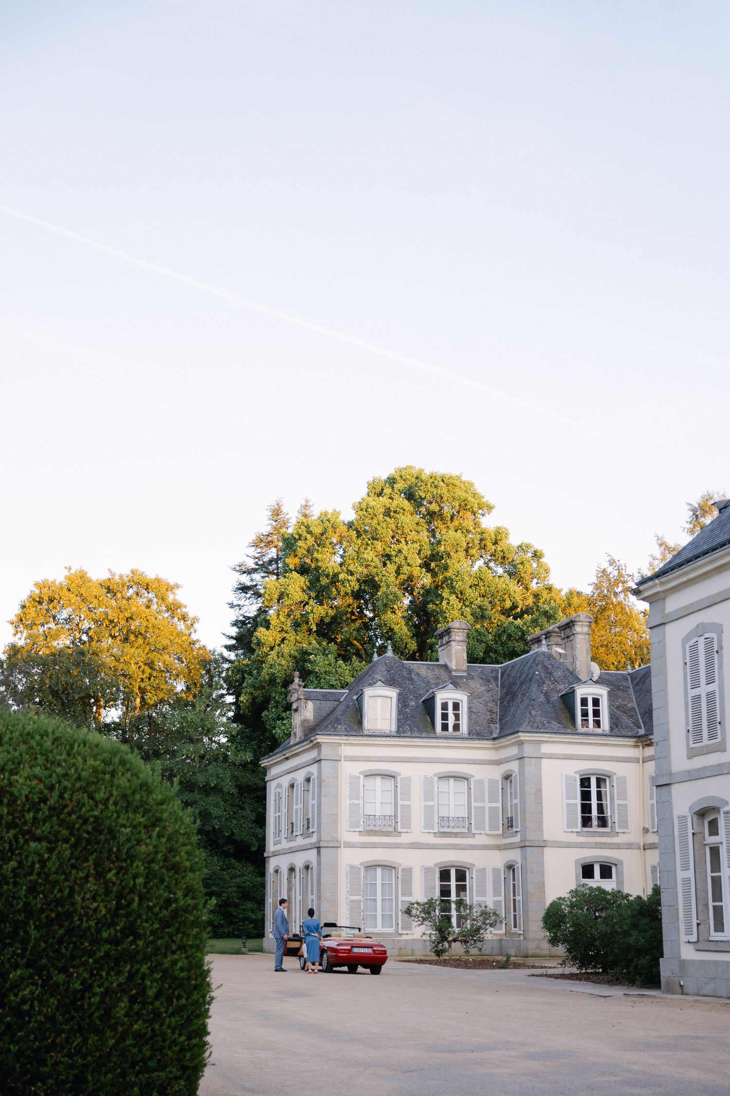 Élégant château français avec des volets gris, des arbres d'automne et une voiture rouge garée dans la cour par une journée claire.