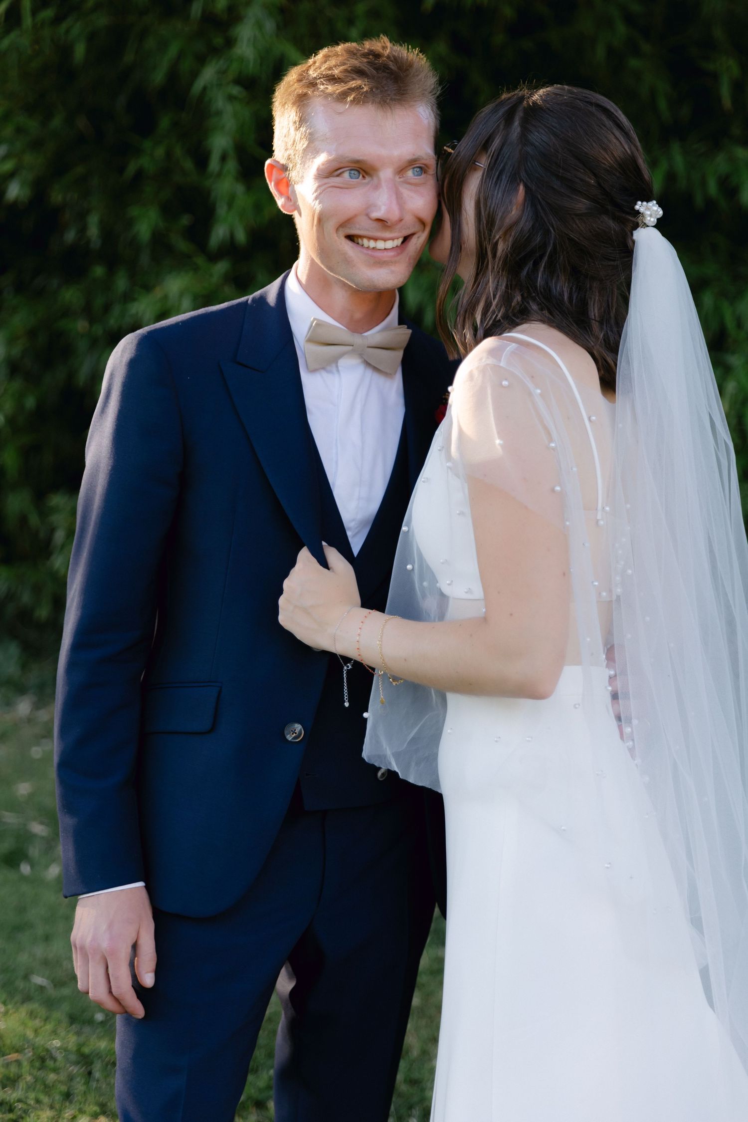 Marié souriant en costume bleu marine et nœud papillon avec mariée en robe de mariée blanche et voile à l'extérieur.