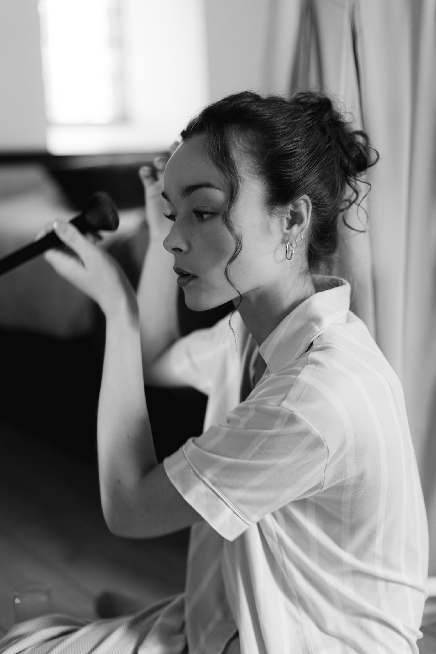 Portrait en noir et blanc d'une jeune femme se maquillant avec un pinceau dans une chemise blanche.