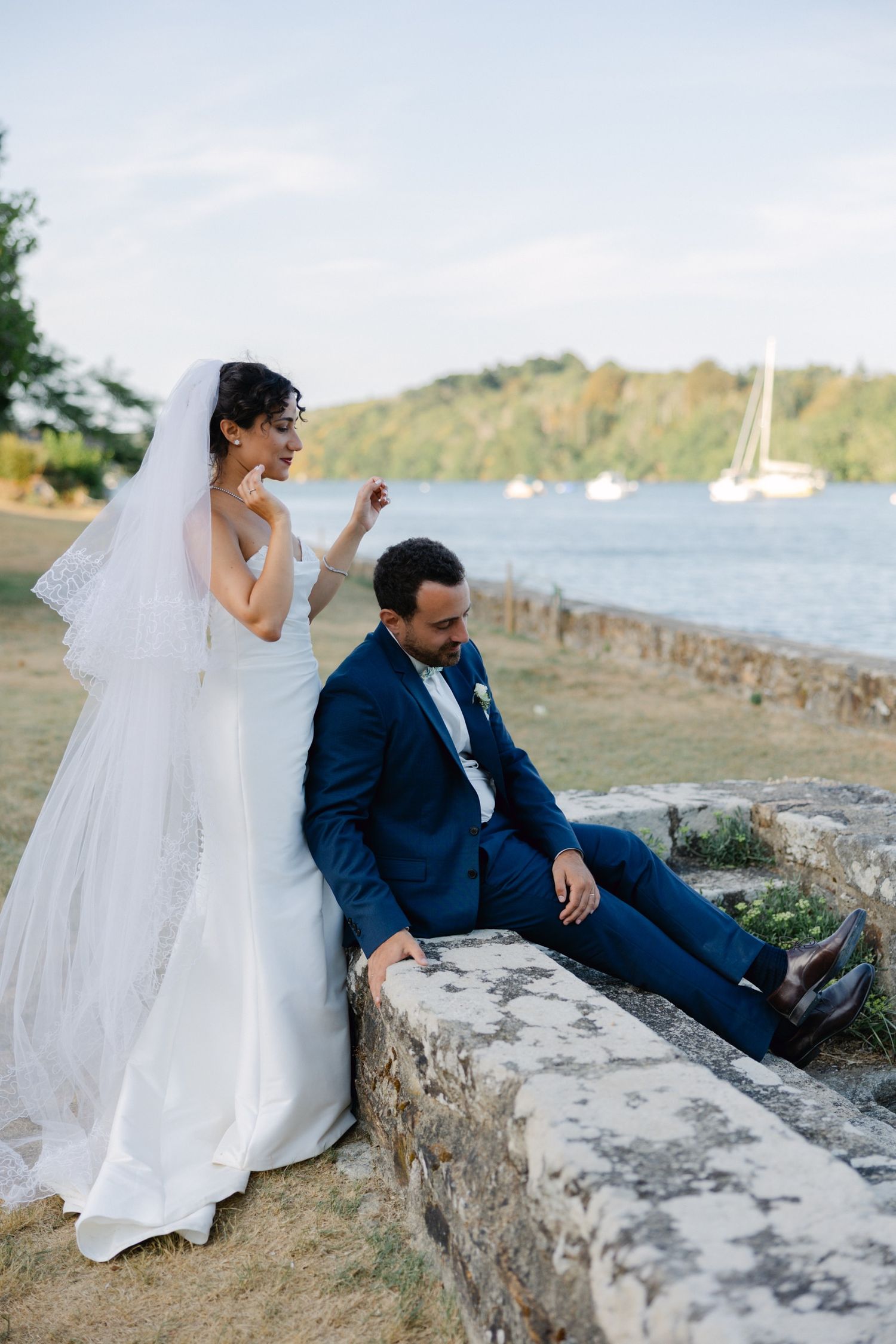 Bride in white gown and veil stands beside groom in navy suit seated on stone wall by waterfront.