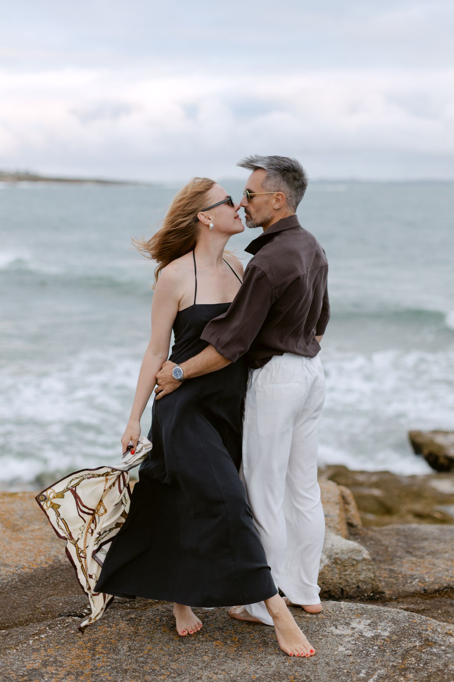 Un couple partage un baiser romantique sur un rivage rocheux, une femme en robe noire tenant une écharpe, un homme en chemise marron.