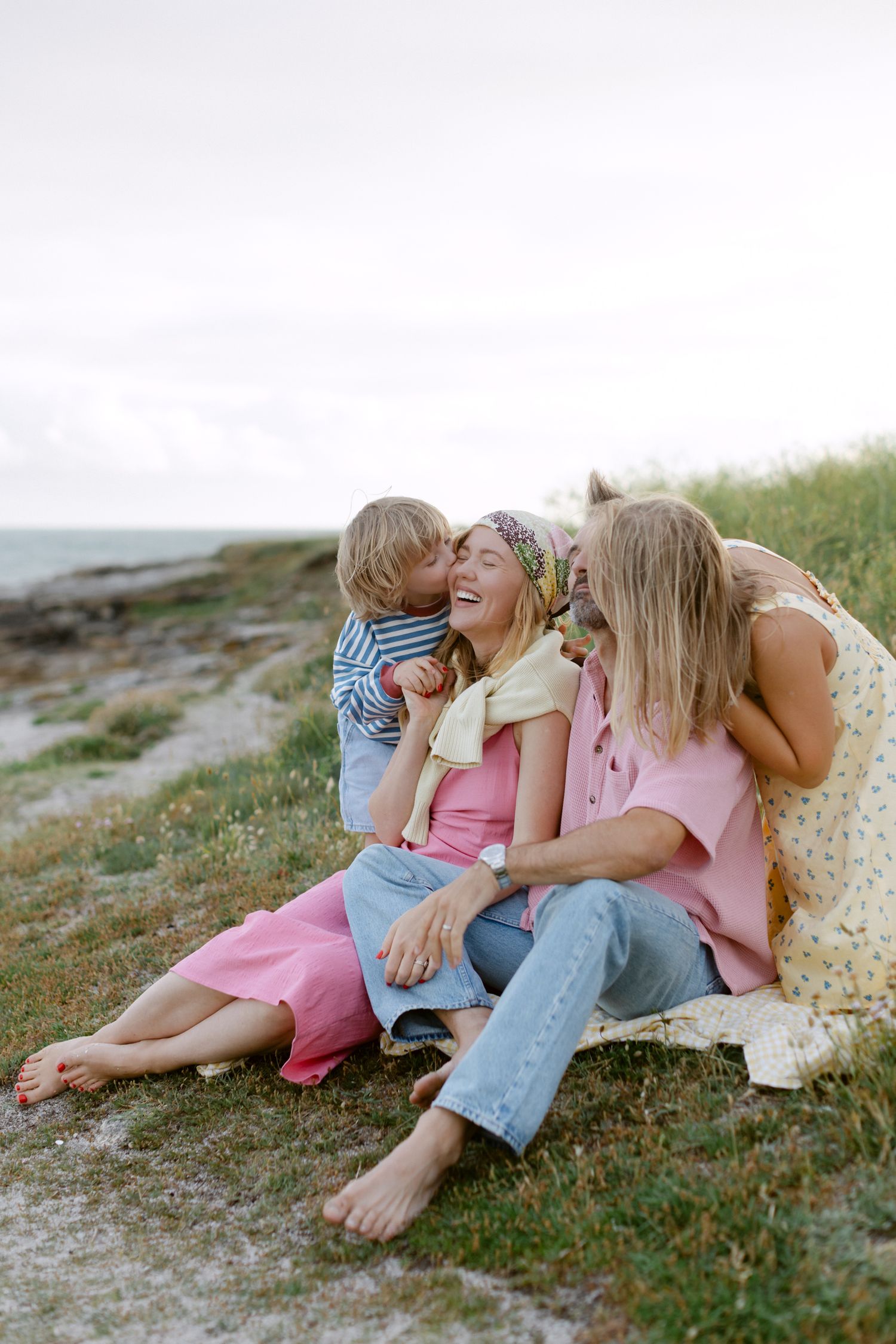 Famille de trois personnes assise sur l'herbe côtière, riant et partageant un baiser, vêtue de tenues d'été décontractées roses et bleues.