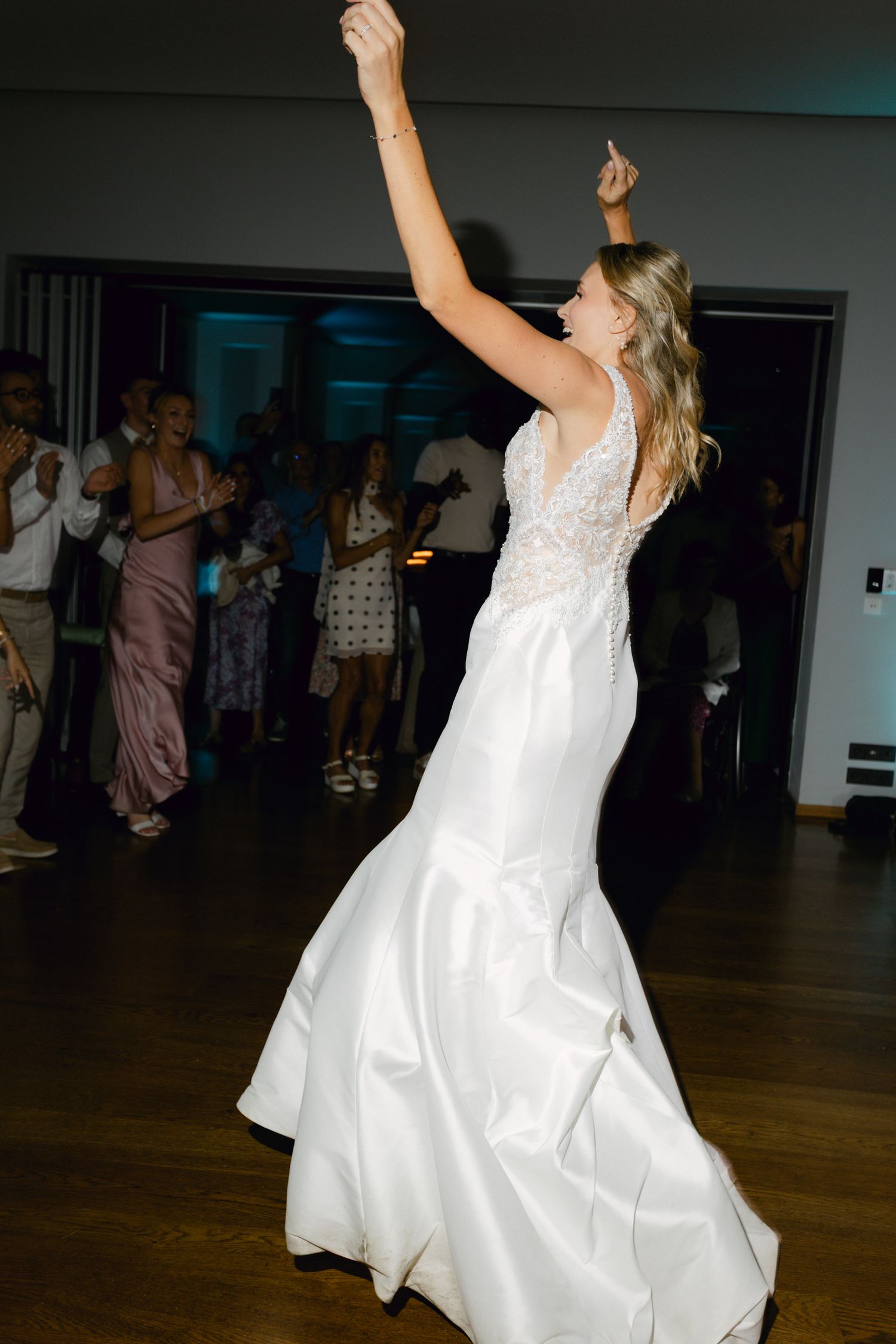 La mariée en robe de sirène en dentelle blanche ajustée lève le bras joyeusement sur la piste de danse de la réception de mariage.