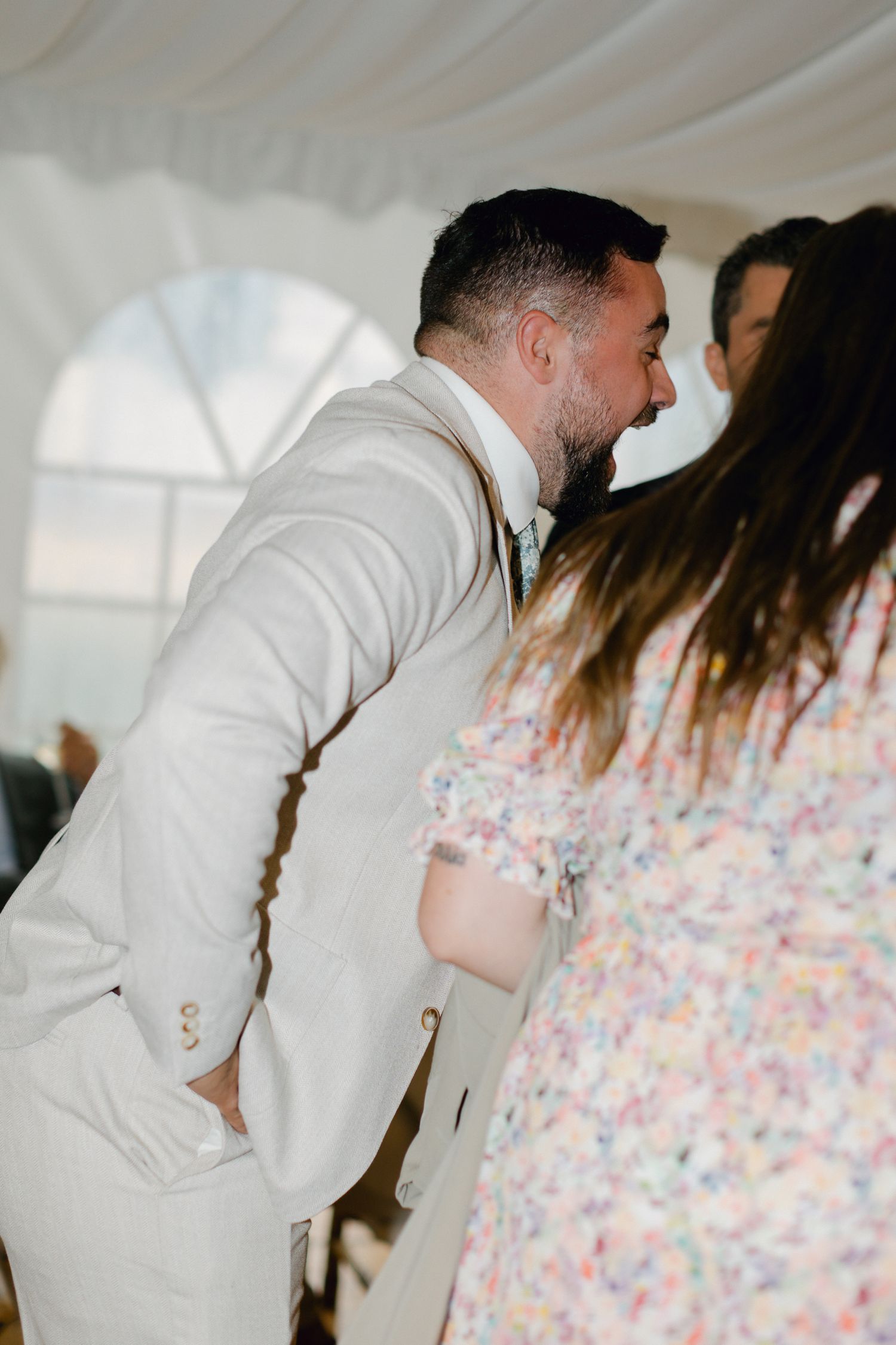 Homme en costume beige accueillant les invités du mariage avec un baiser dans une tente blanche.
