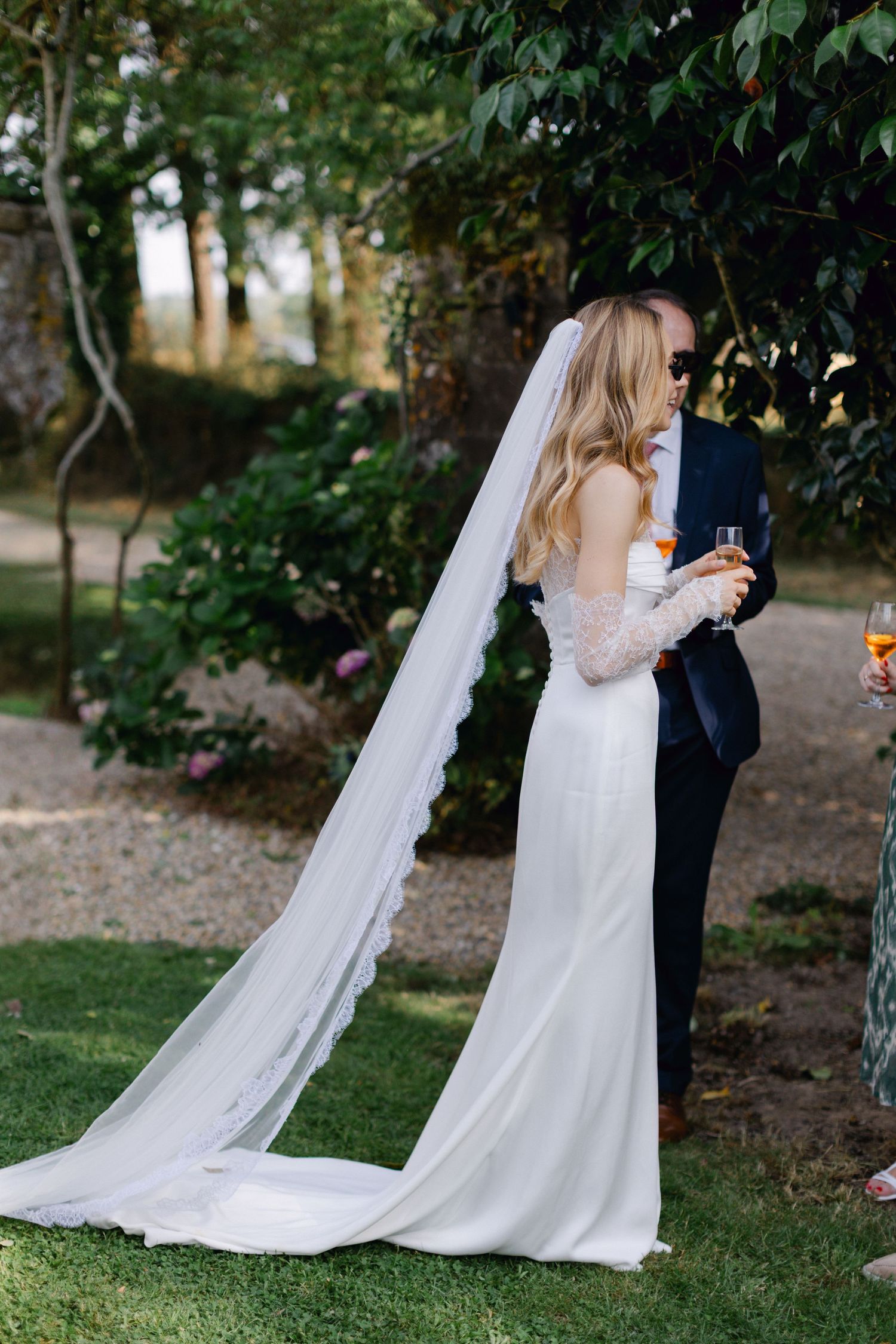 Mariée en robe blanche avec un long voile garni de dentelle debout avec un invité tenant des verres à champagne.