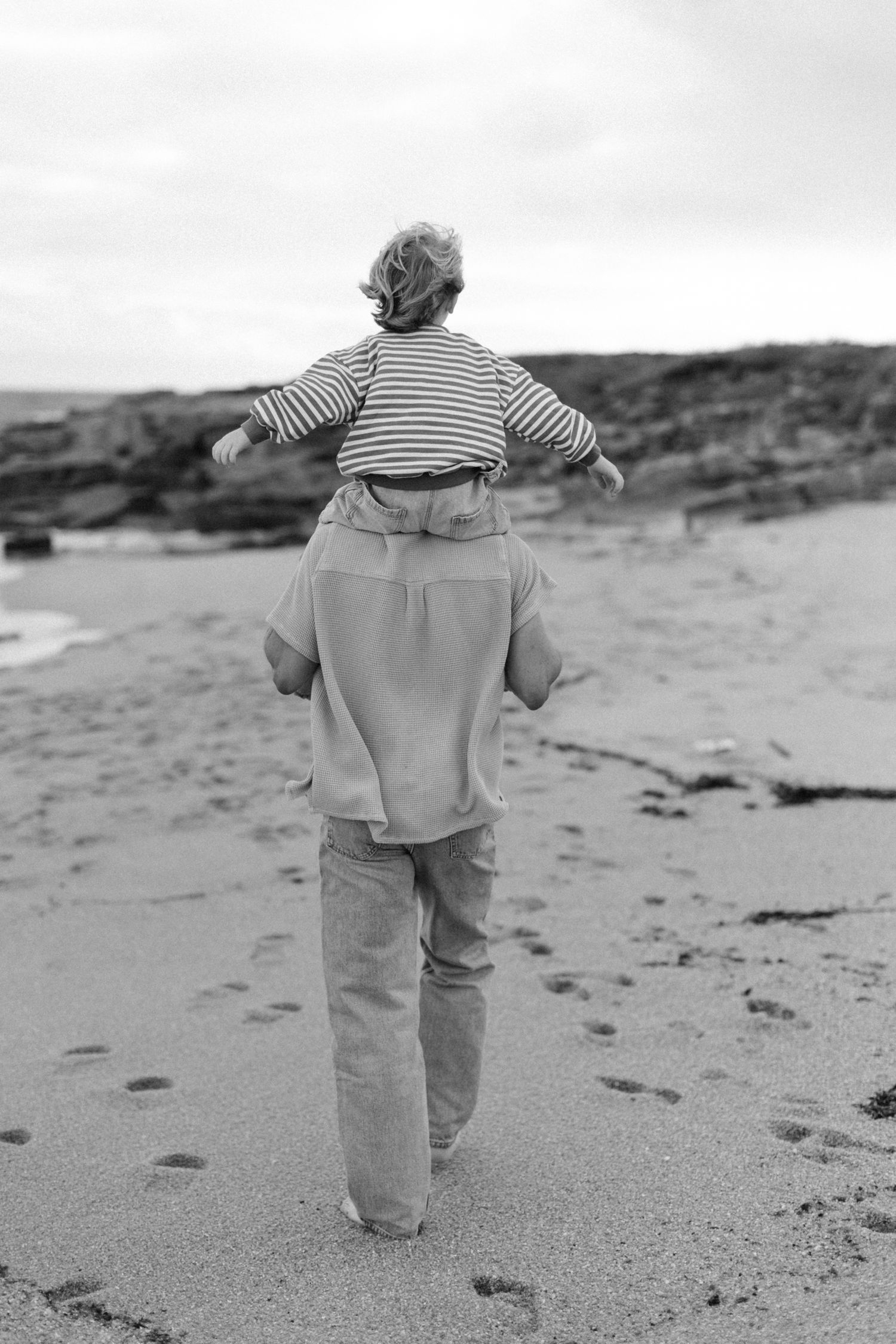 Photo en noir et blanc d'un adulte portant un enfant sur les épaules marchant le long de la plage de sable avec un littoral rocheux.