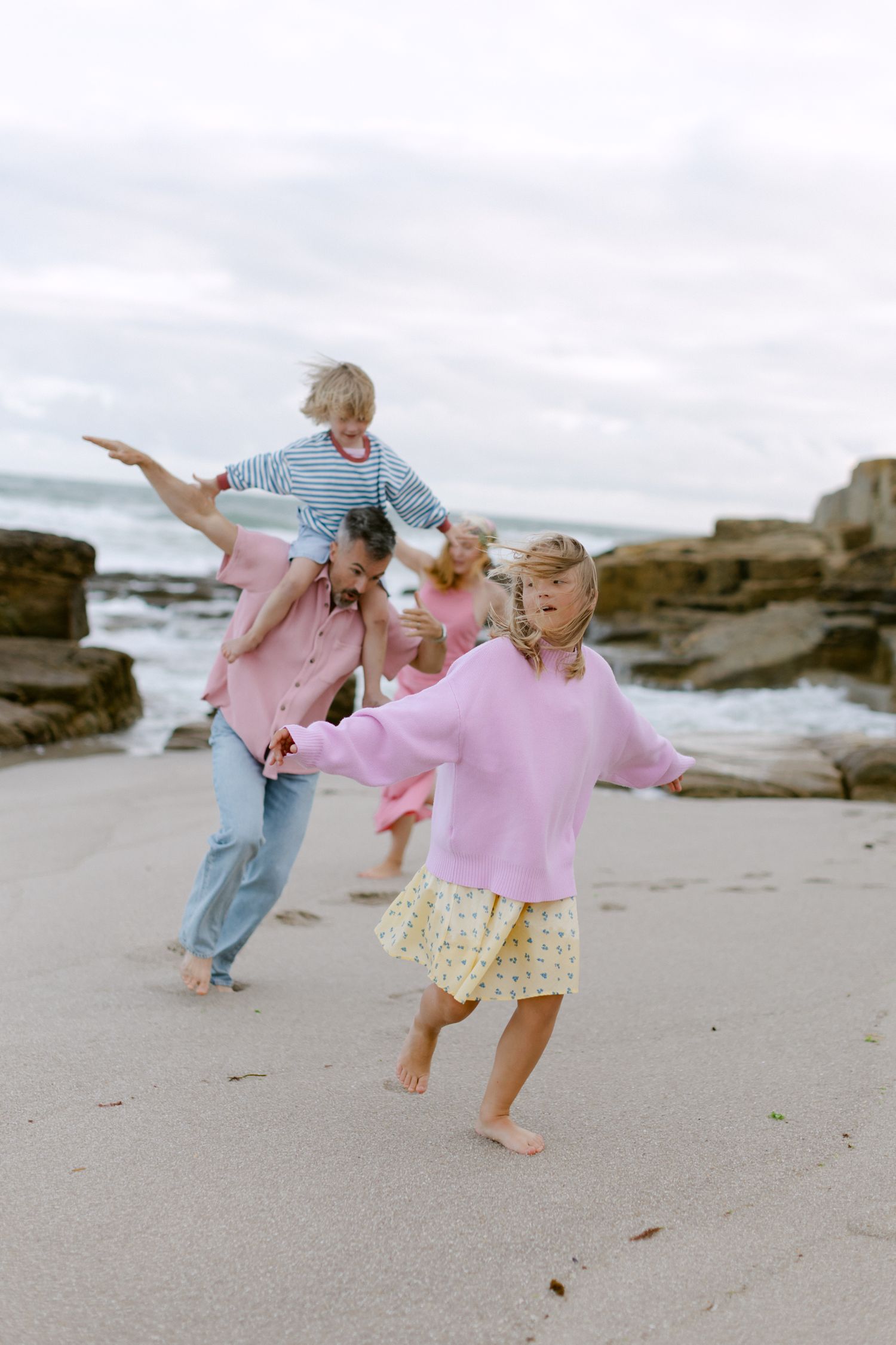 Une famille joue joyeusement sur une plage de sable, avec des enfants en pulls roses et un parent faisant un tour à cheval près des rivages