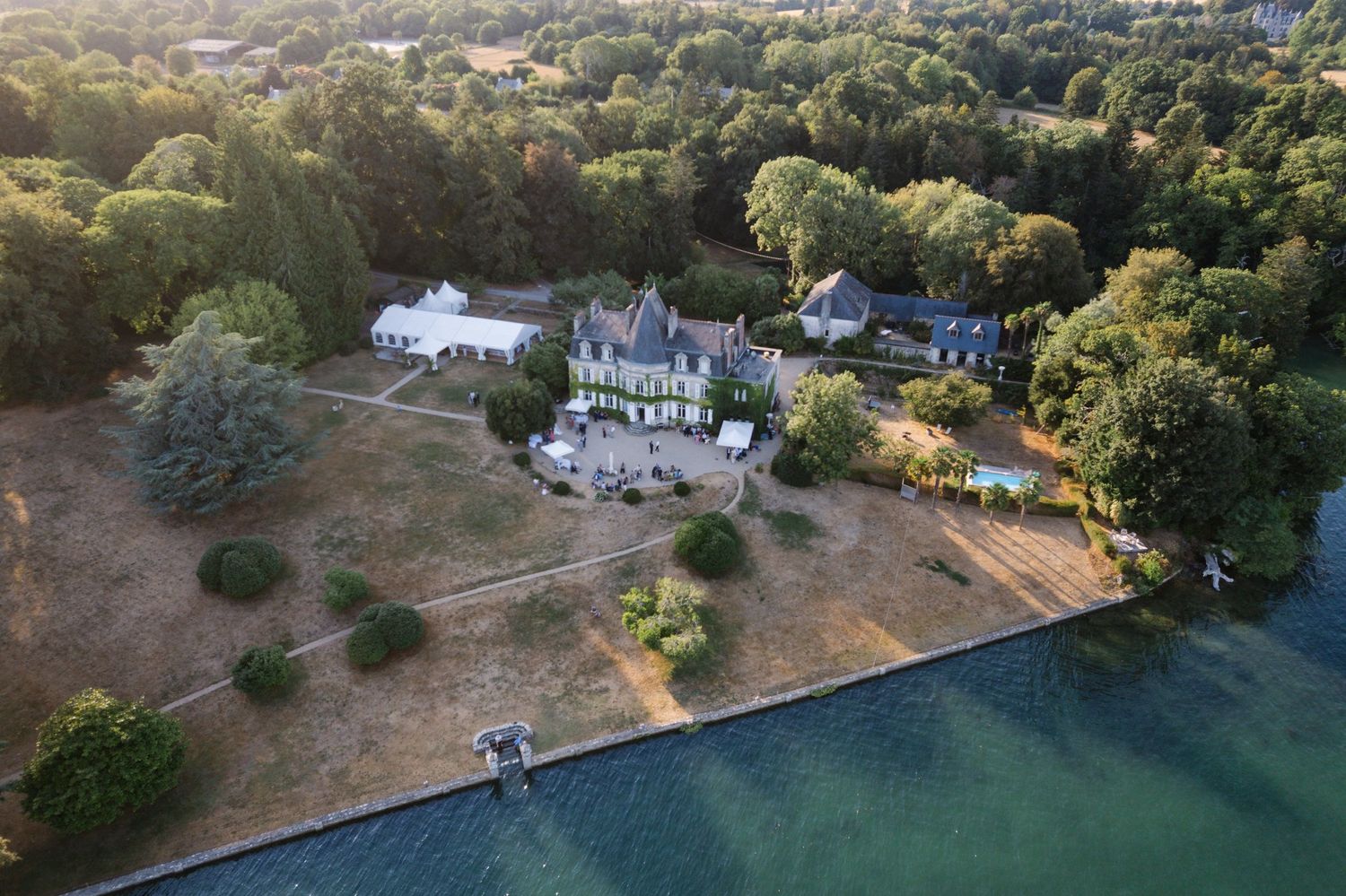 Aerial view of a grand estate with a chateau surrounded by lush trees along a serene waterfront.