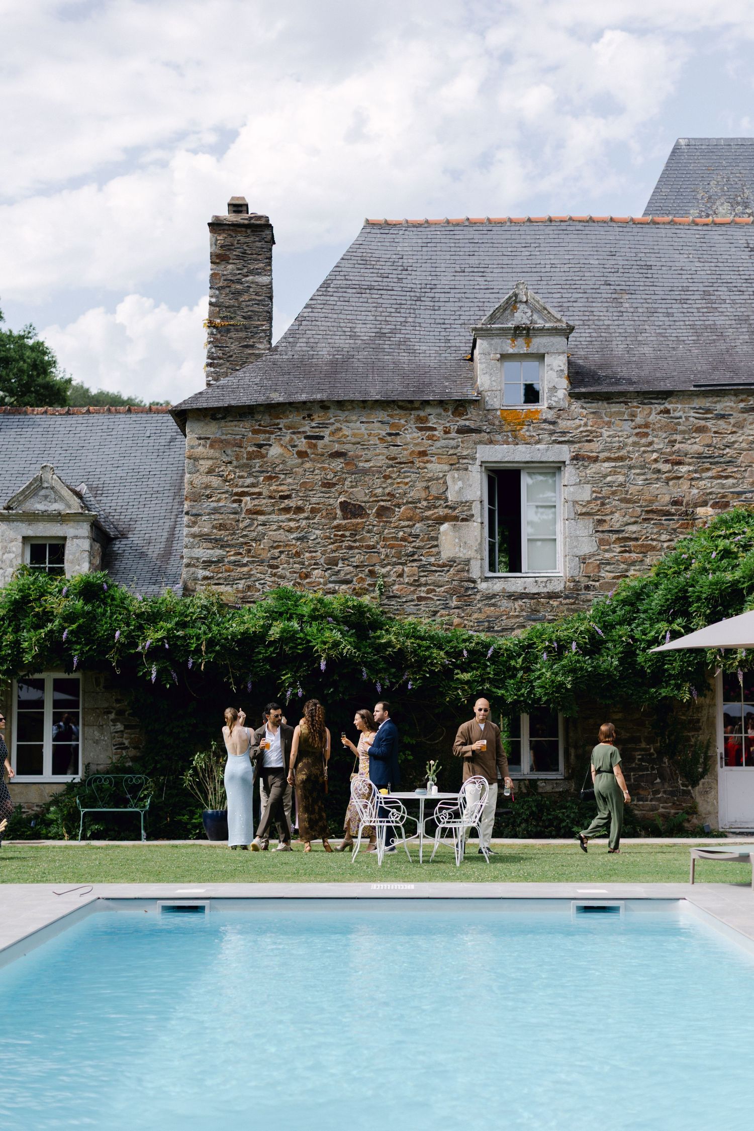 Les invités au mariage socialisent à côté d'une piscine dans un manoir en pierre rustique couvert de vignes grimpantes.