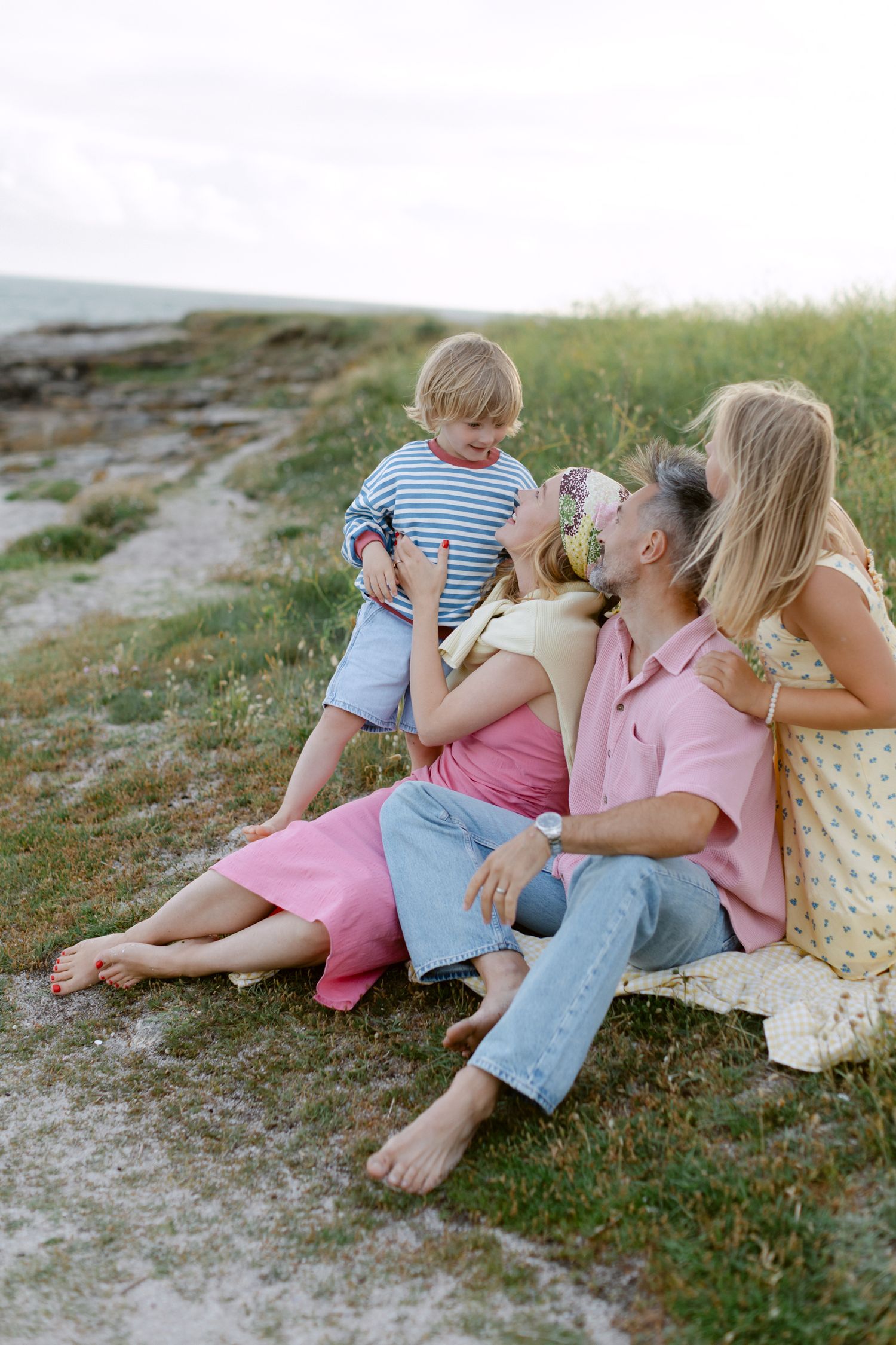 Famille assise sur un chemin côtier herbeux, parents se relaxant tandis que le jeune garçon en chemise rayée se tient à proximité.