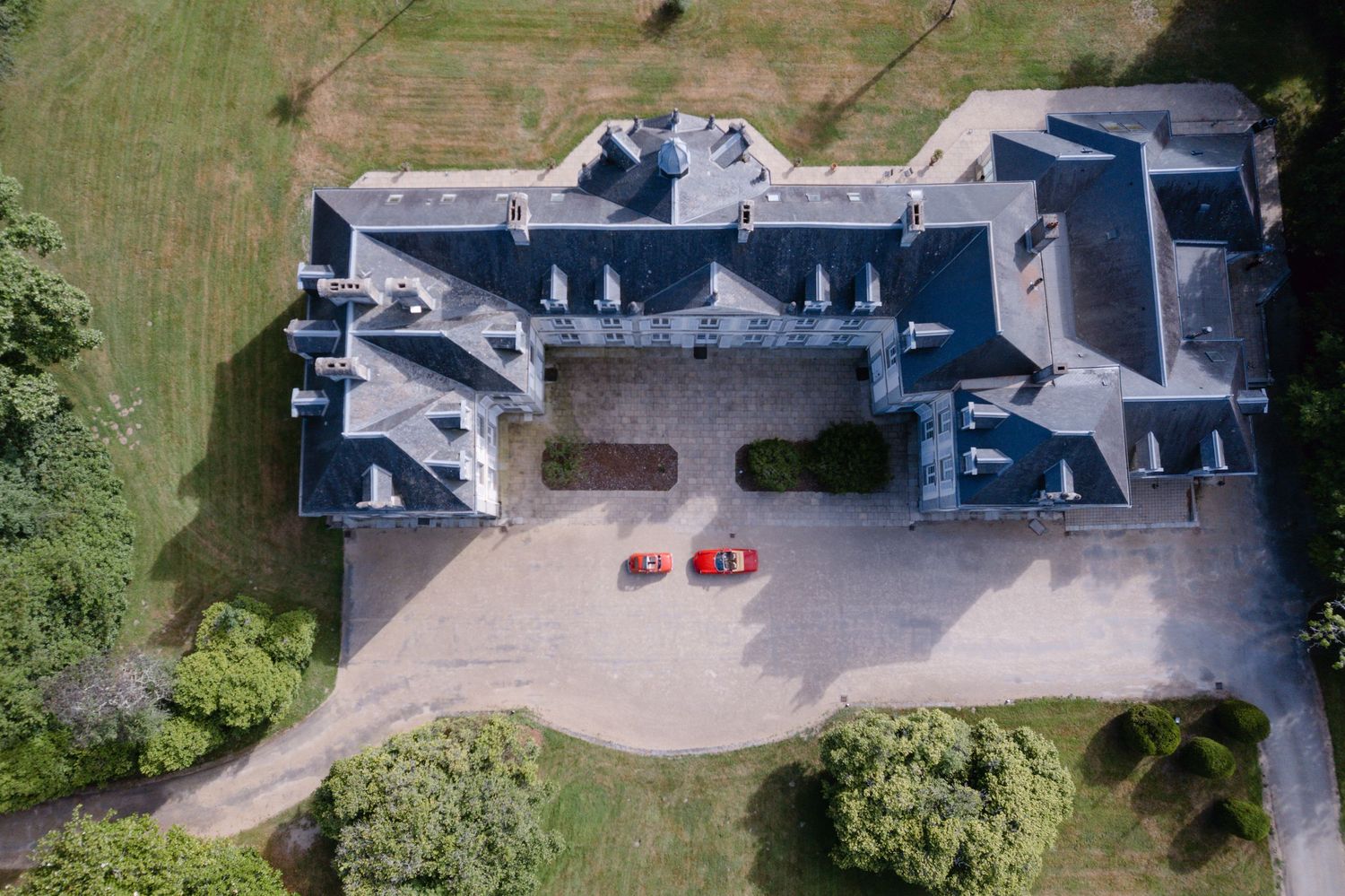 Vue aérienne d'un grand manoir en pierre avec deux voitures rouges garées dans la cour, entourée d'un terrain verdoyant.