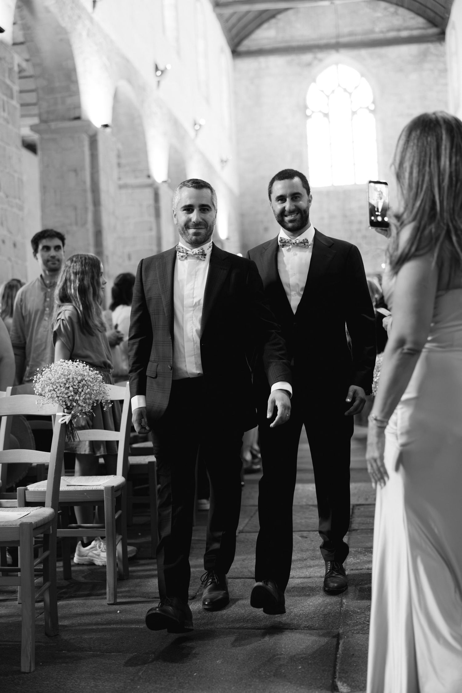 Two grooms in suits and bow ties walk down church aisle smiling on their wedding day.