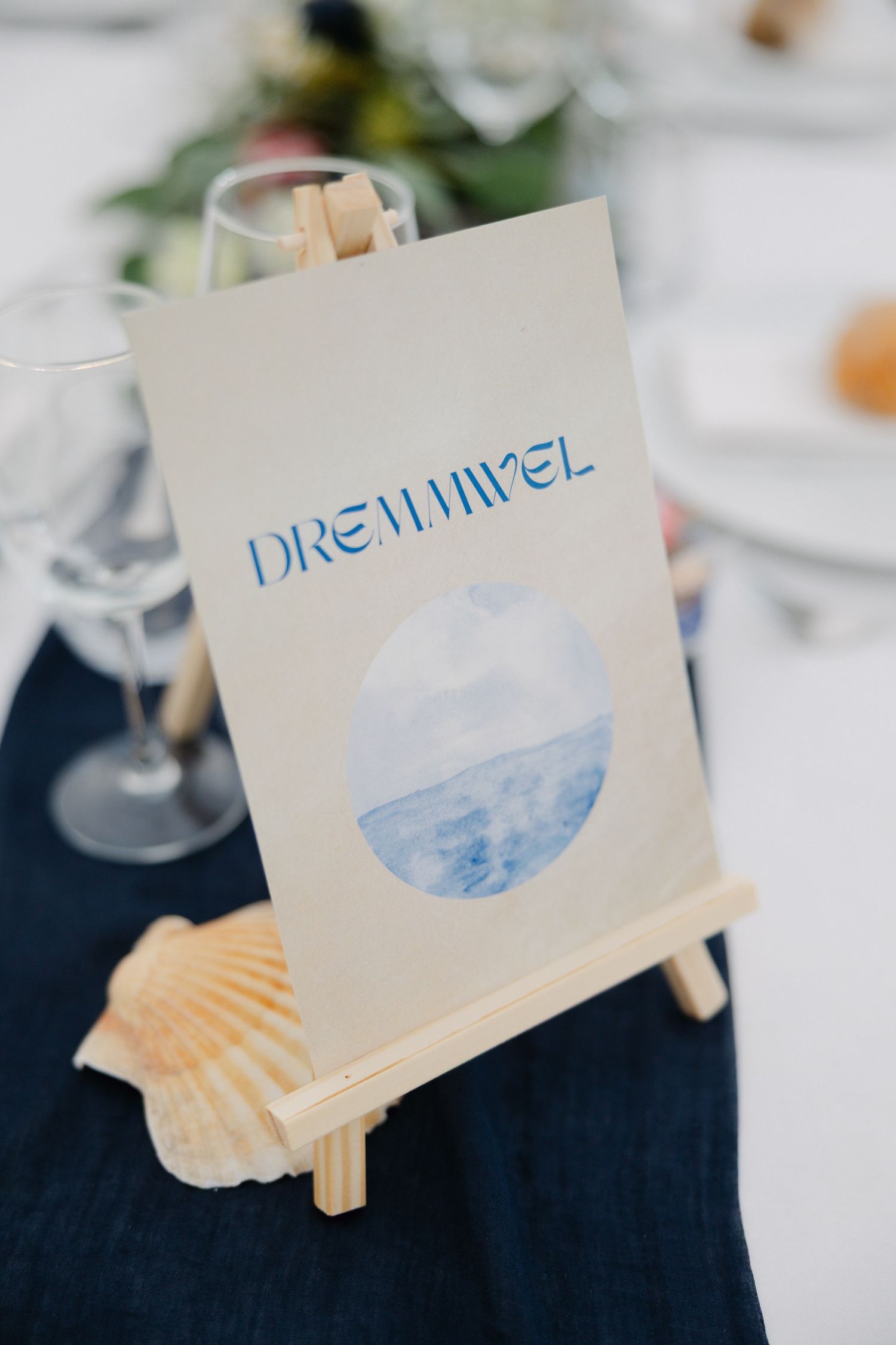 A small easel displays a beach-themed wedding table card reading 'Dreamivel' with a circular ocean watercolor print.