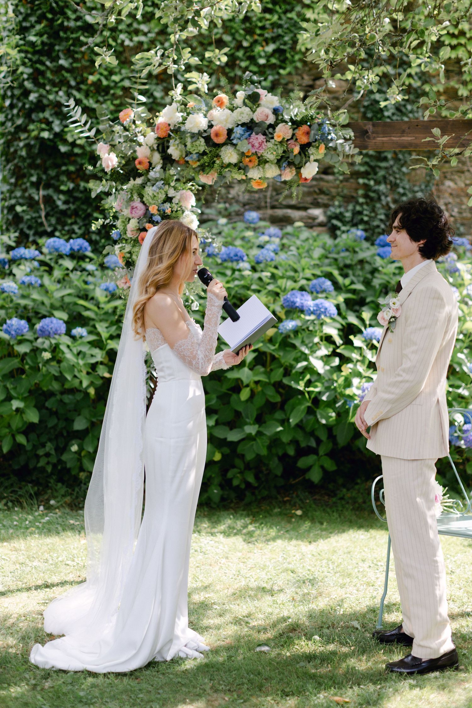 La mariée lit des vœux lors d'une cérémonie de mariage dans un jardin en plein air sous une arche florale colorée avec des hortensias en arr