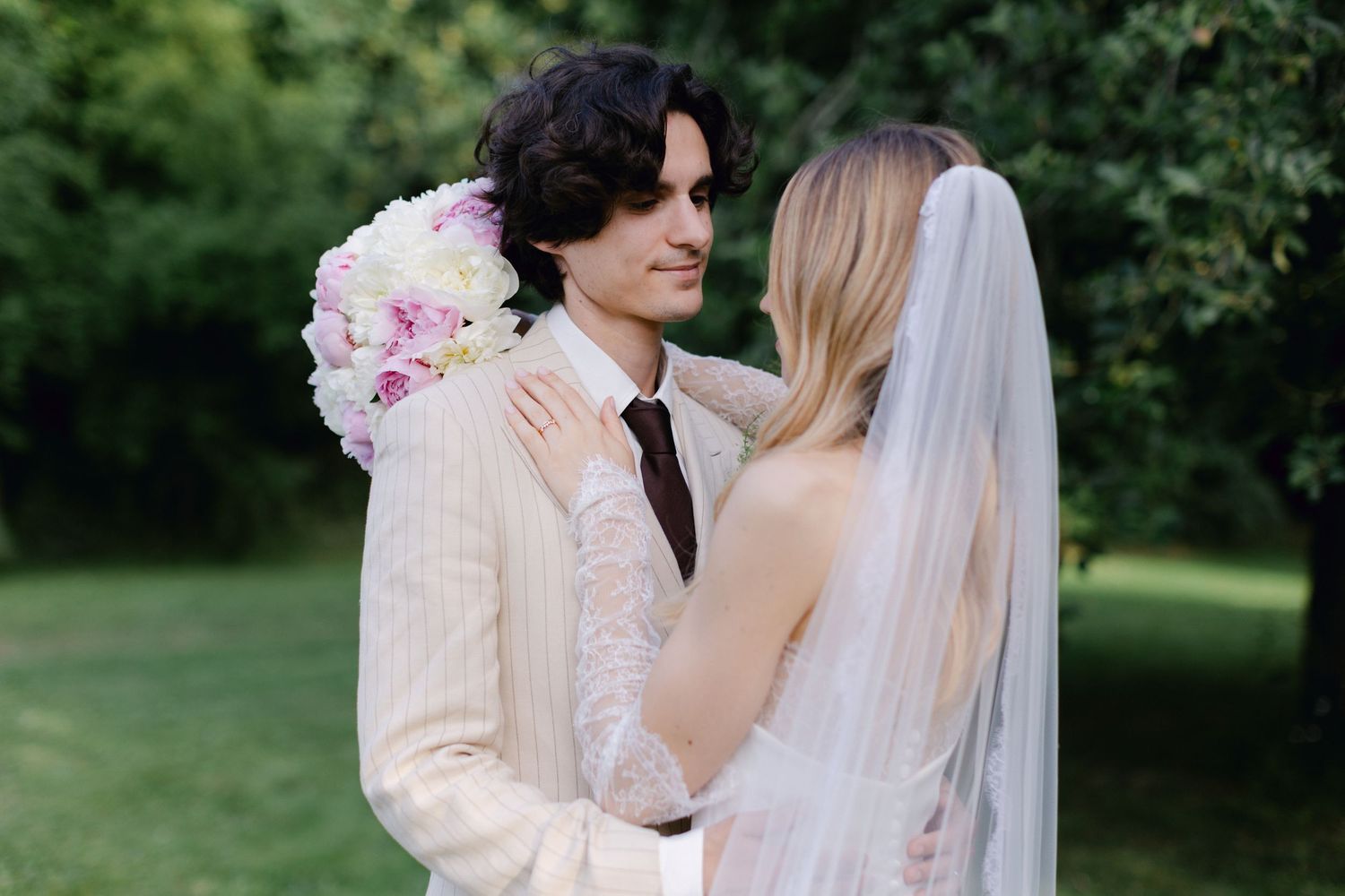 Le marié en costume à rayures beiges tient un bouquet floral rose tandis que la mariée en robe blanche à manches en dentelle l'embrasse.