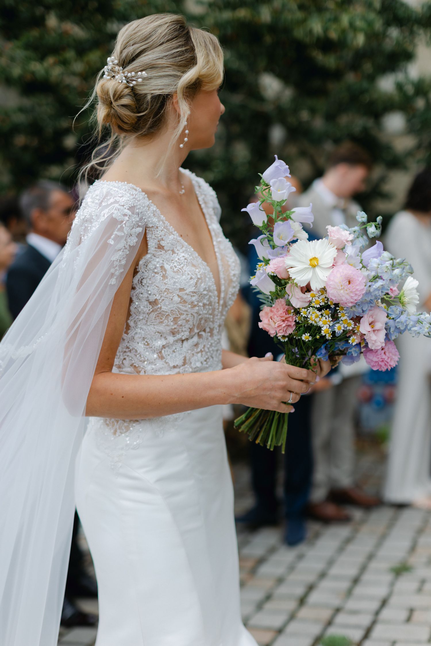 La mariée blonde en robe cape en dentelle tient un bouquet pastel de fleurs roses, bleues et blanches à l'extérieur.