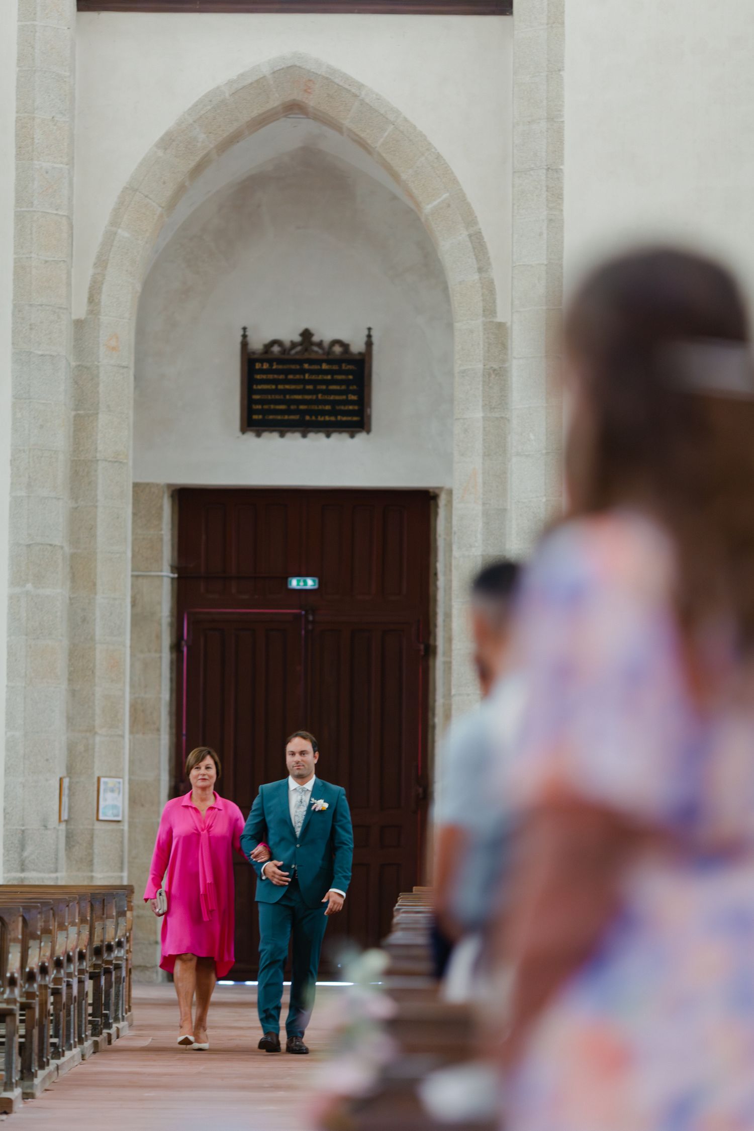 Le marié en costume sarcelle accompagne une femme en robe rose dans l'allée de l'église avant le début de la cérémonie de mariage.