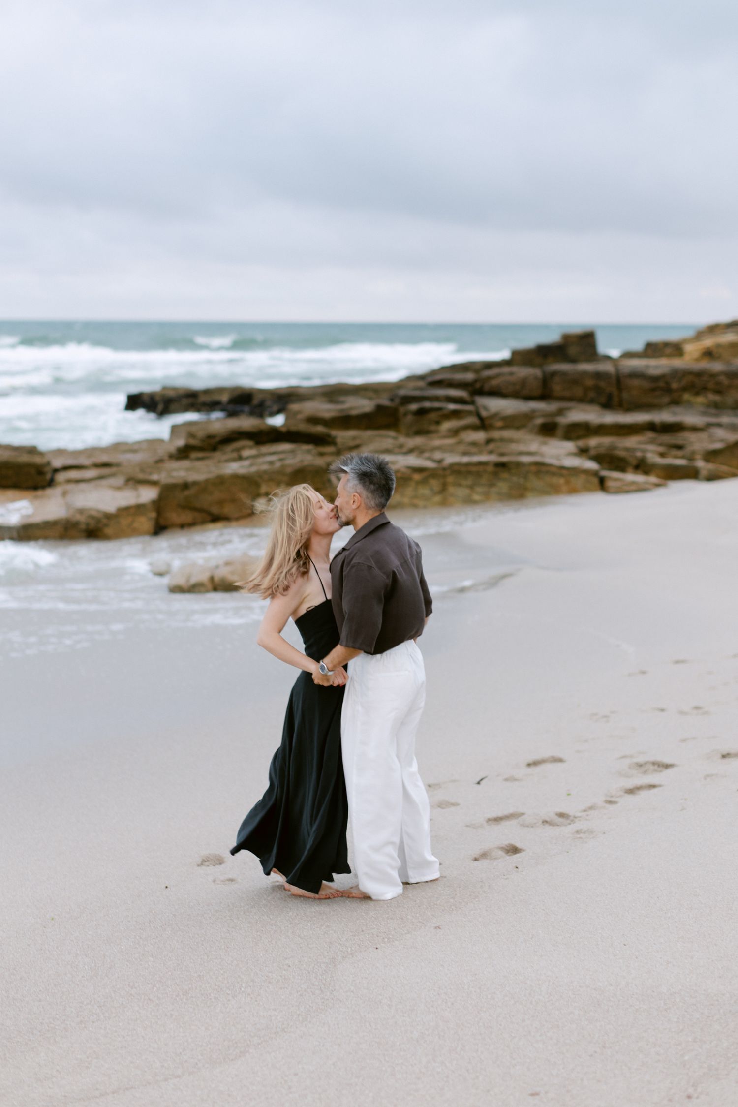 Couple romantique s'embrassant sur une plage de sable avec un rivage rocheux et des vagues de l'océan par une journée nuageuse.