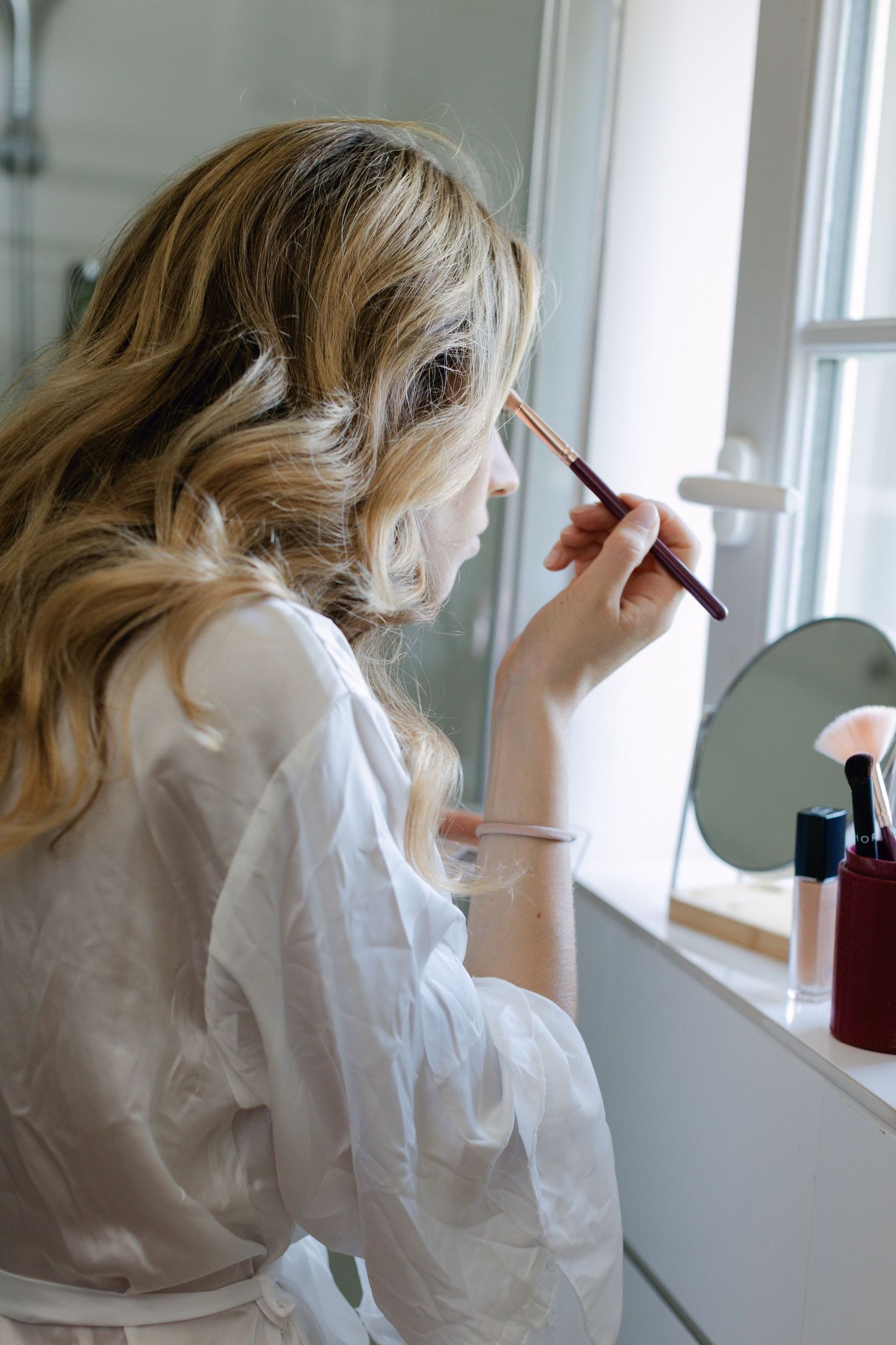 Mariée blonde appliquant du fard à paupières avec un pinceau de maquillage tout en se préparant dans une vanité de salle de bain lumineuse.