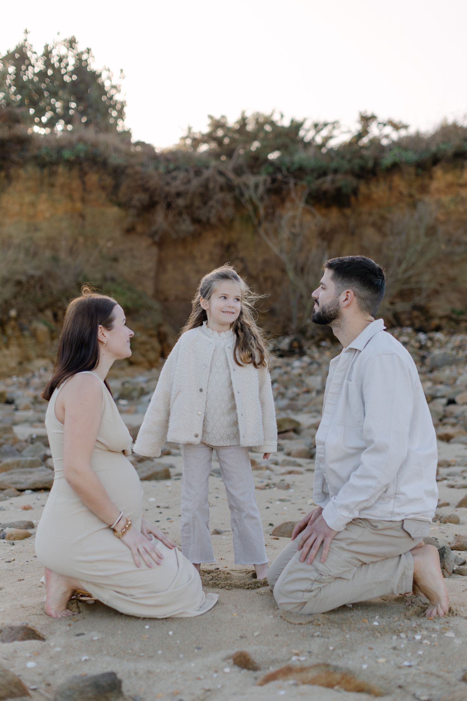 Portrait de famille en attente sur une plage rocheuse avec des parents agenouillés à côté d'une fille souriante dans des tons neutres.
