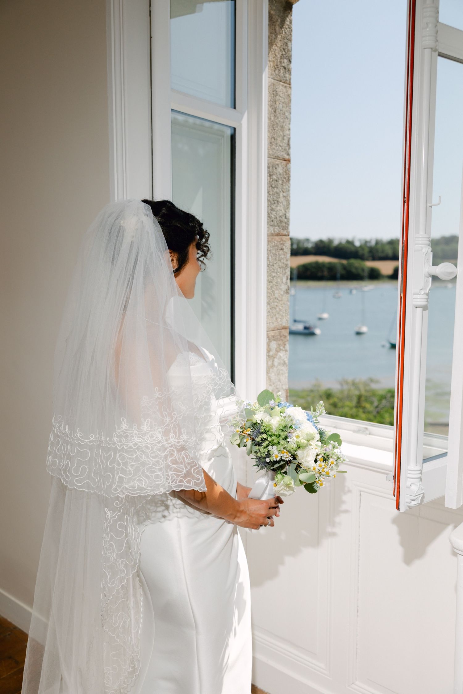 Bride in lace veil holding white bouquet gazes out window at serene waterway with sailboats on sunny wedding day.