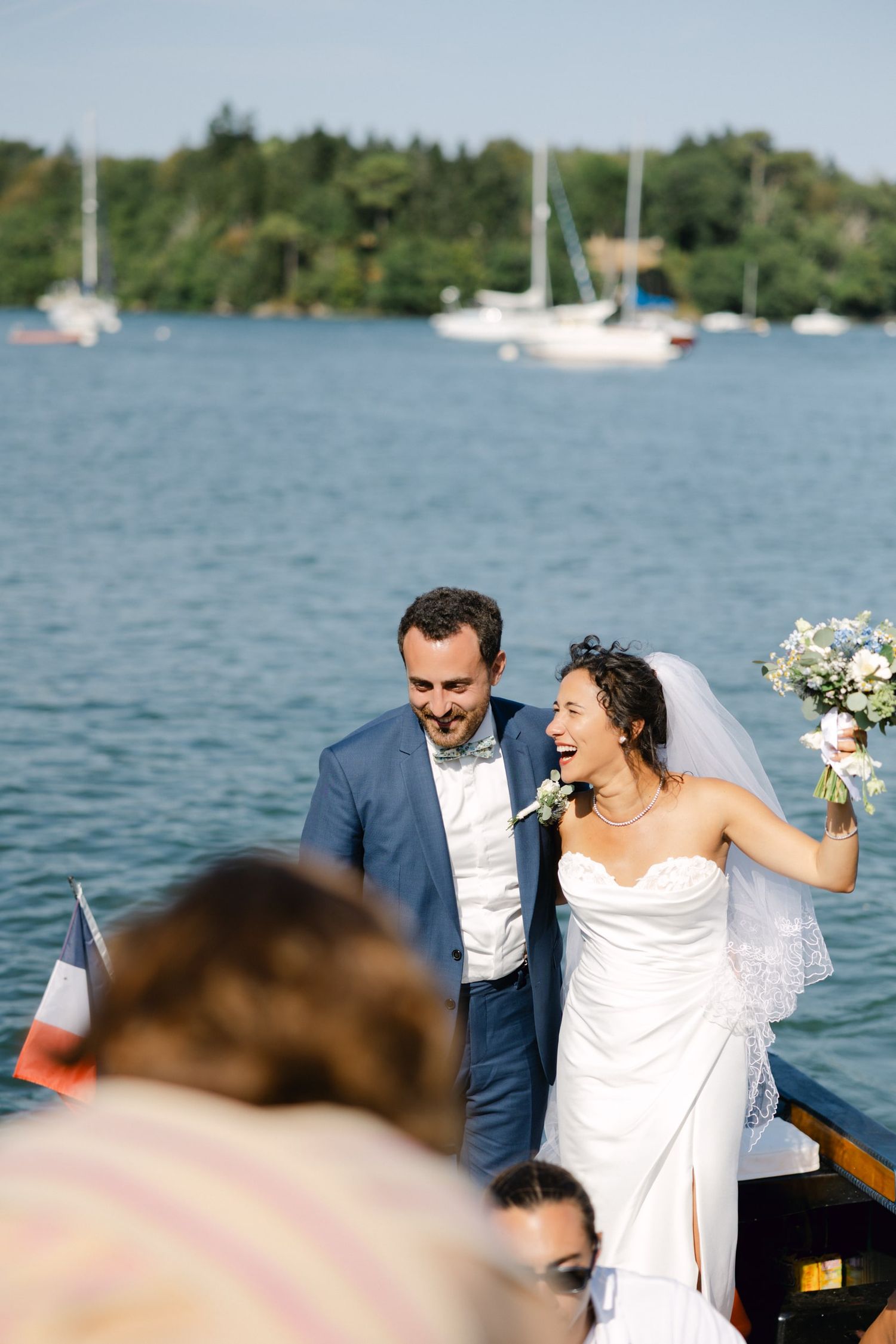 Happy newlyweds laughing on a boat with a lake and sailboats in the background on their wedding day.