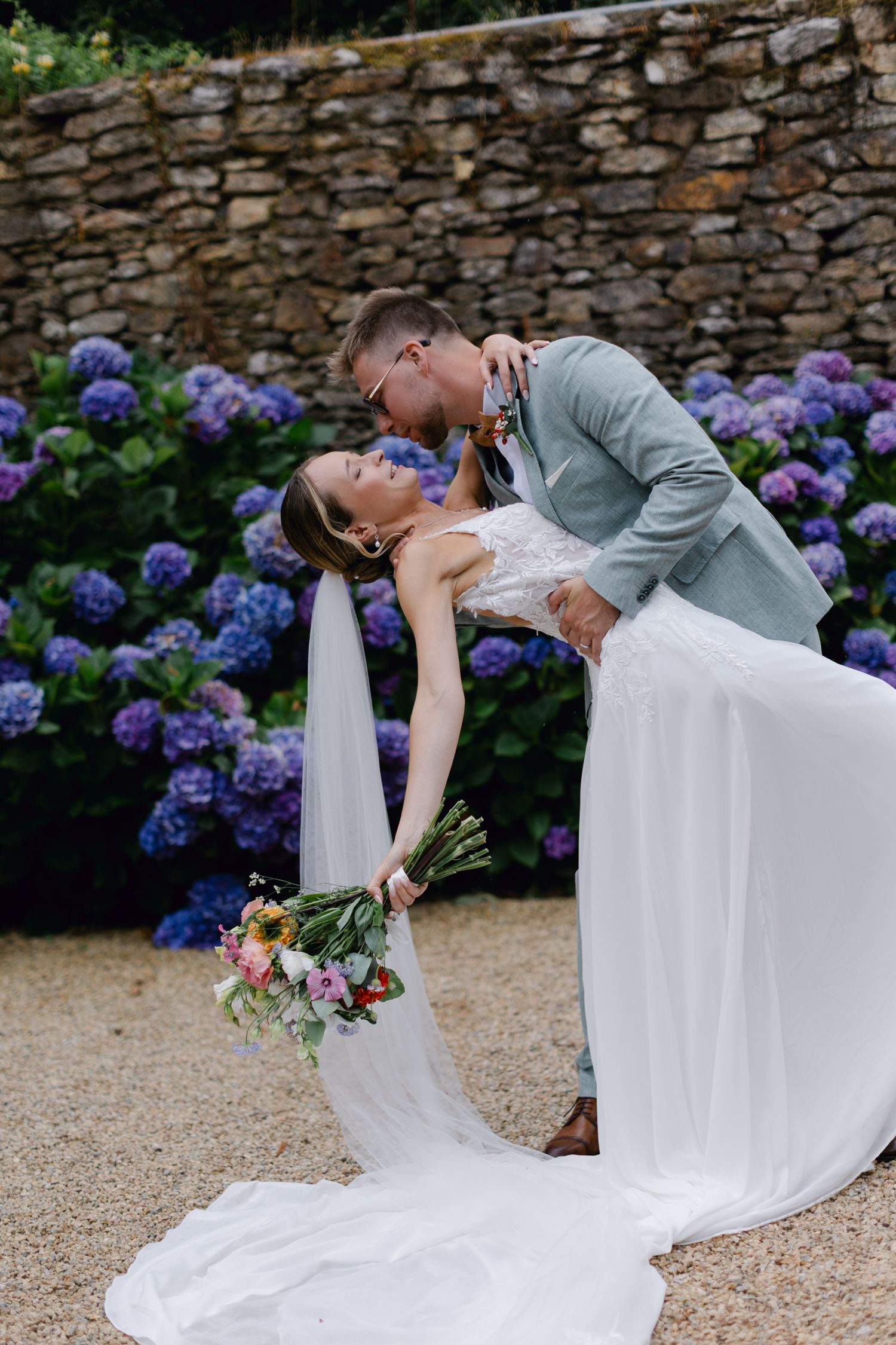 Le marié plonge la mariée pour un baiser romantique devant des hortensias violets en fleurs lors d'un mariage en plein air.