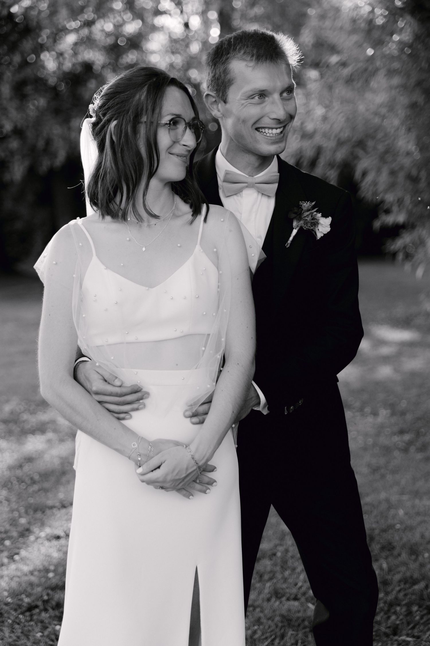Photo de mariage en noir et blanc d'une mariée souriante en robe blanche et d'un marié en smoking avec nœud papillon embrassant à l'extérieu