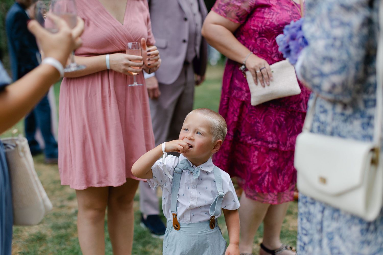 Adorable jeune garçon en bretelles et nœud papillon buvant lors d'une réception de mariage en plein air parmi les invités.