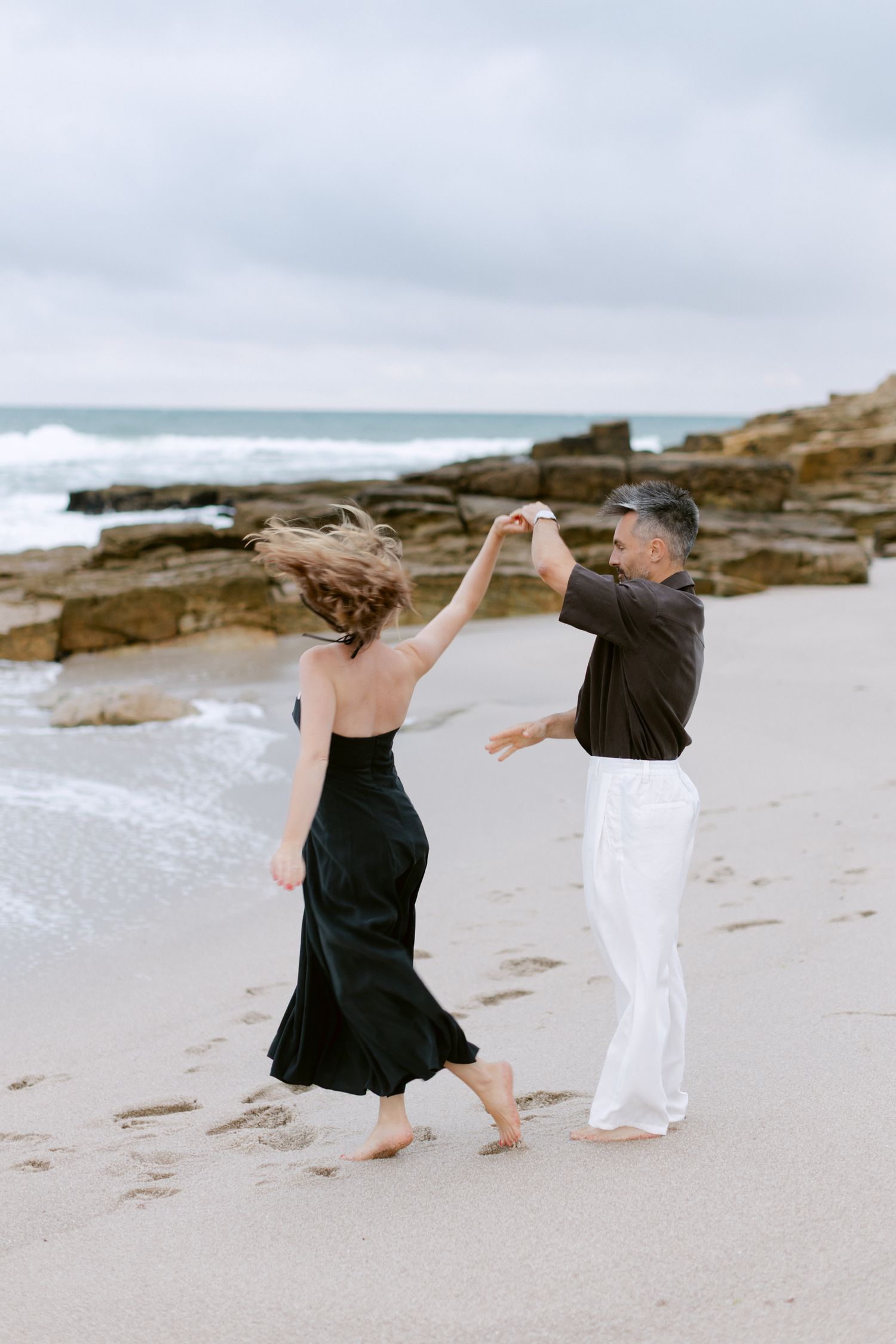 Couple dansant pieds nus sur la plage de sable, femme en robe noire, homme en pantalon blanc, vagues de l'océan et rochers derrière eux.