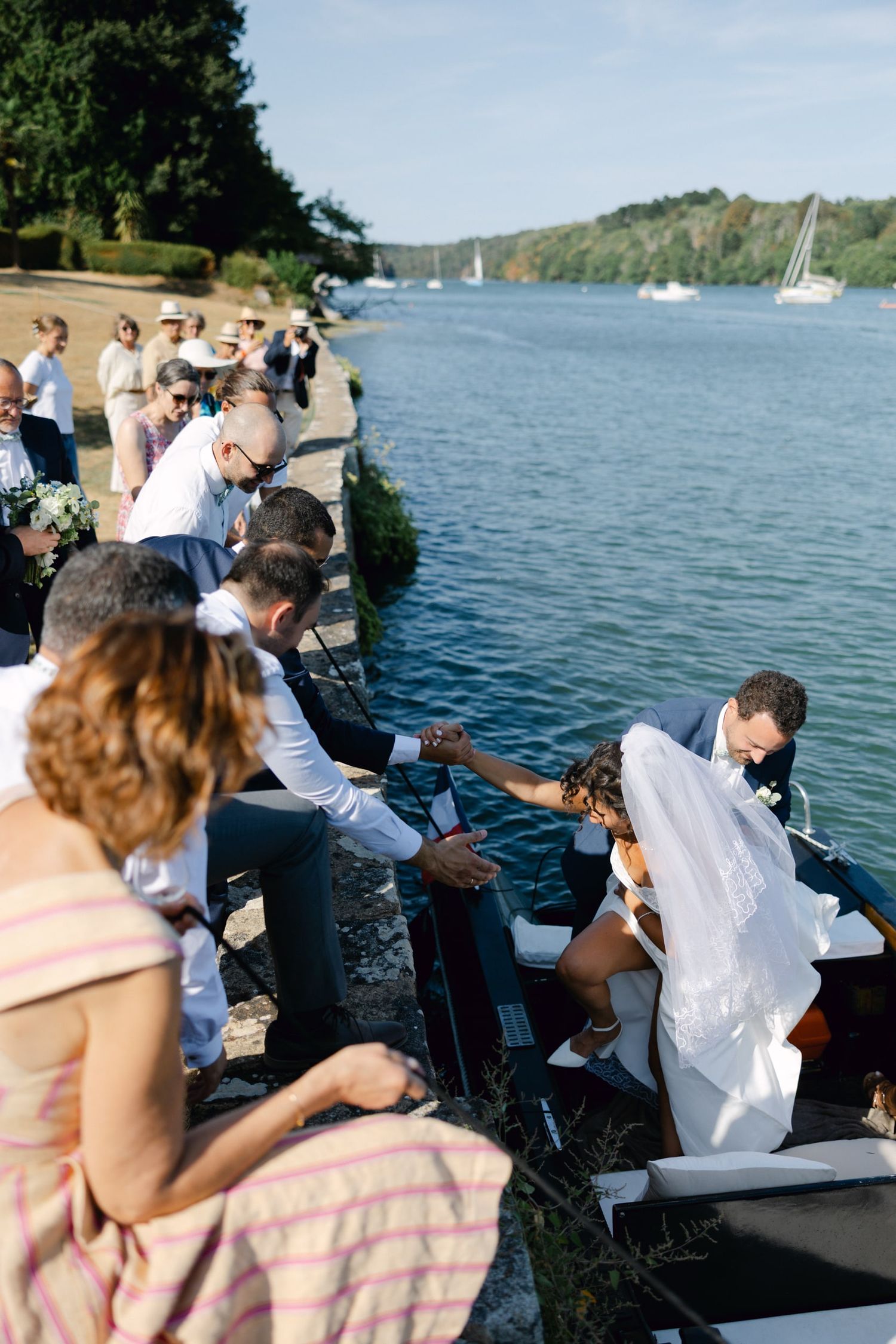 Bride and groom boarding a boat by the waterside, assisted by wedding guests on a sunny day.