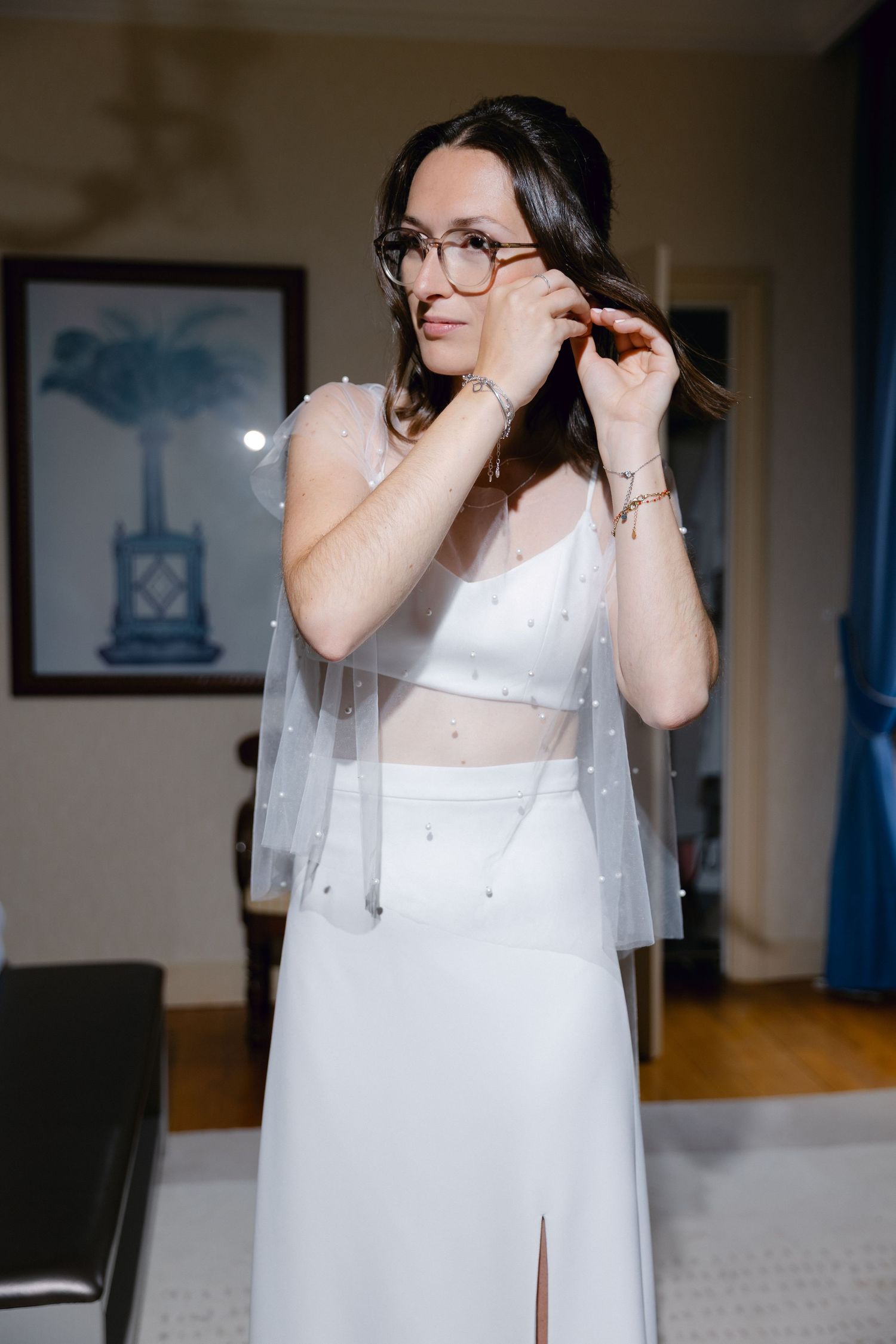 Mariée en robe de mariée blanche deux pièces et voile avec des perles ajustant ses lunettes dans une pièce lumineuse.