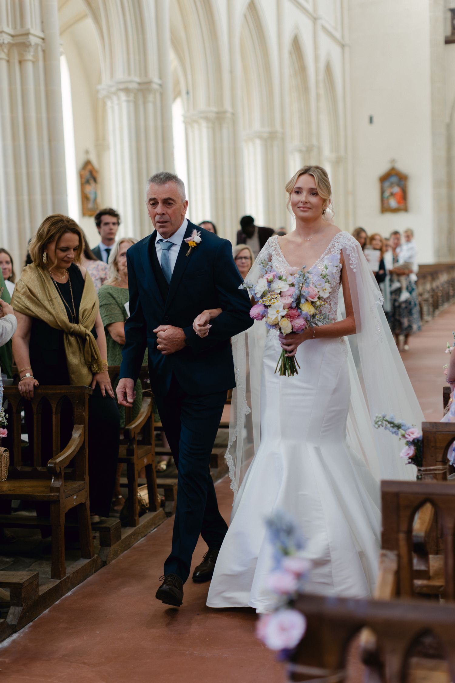 La mariée portant un bouquet coloré marche dans l'allée de l'église avec son père en costume bleu marine lors de la cérémonie de mariage.