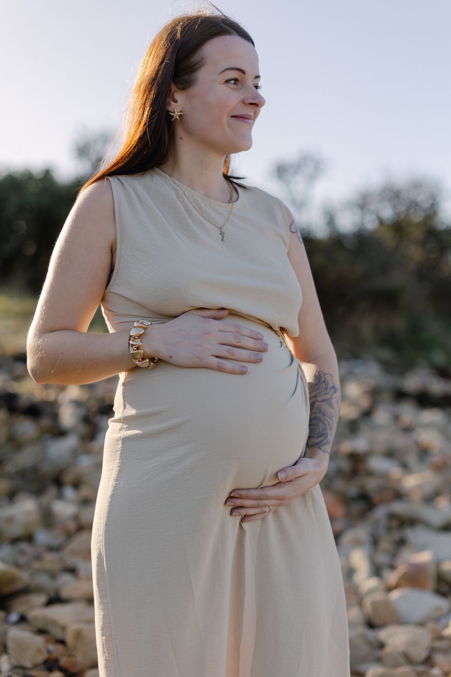 Femme enceinte souriante en robe beige sans manches tenant la bosse de bébé à l'extérieur au soleil doré.