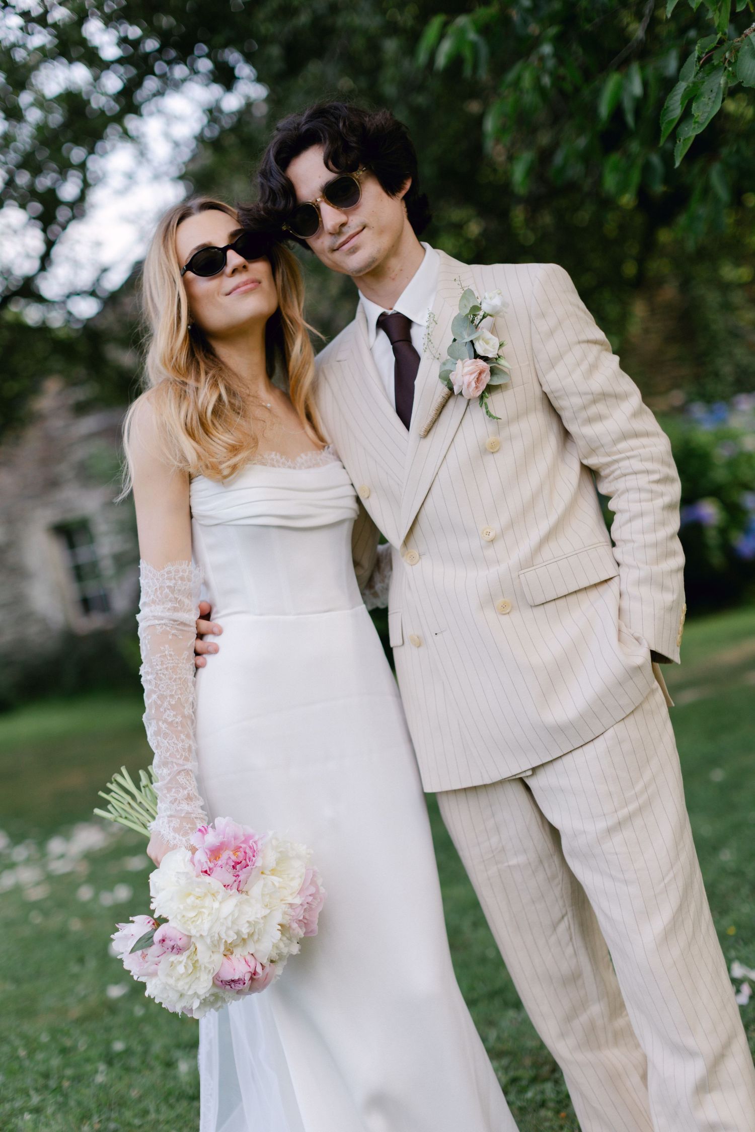 Un couple élégant en lunettes de soleil pose lors d'un mariage en plein air, mariée en robe blanche avec bouquet rose, marié en costume.