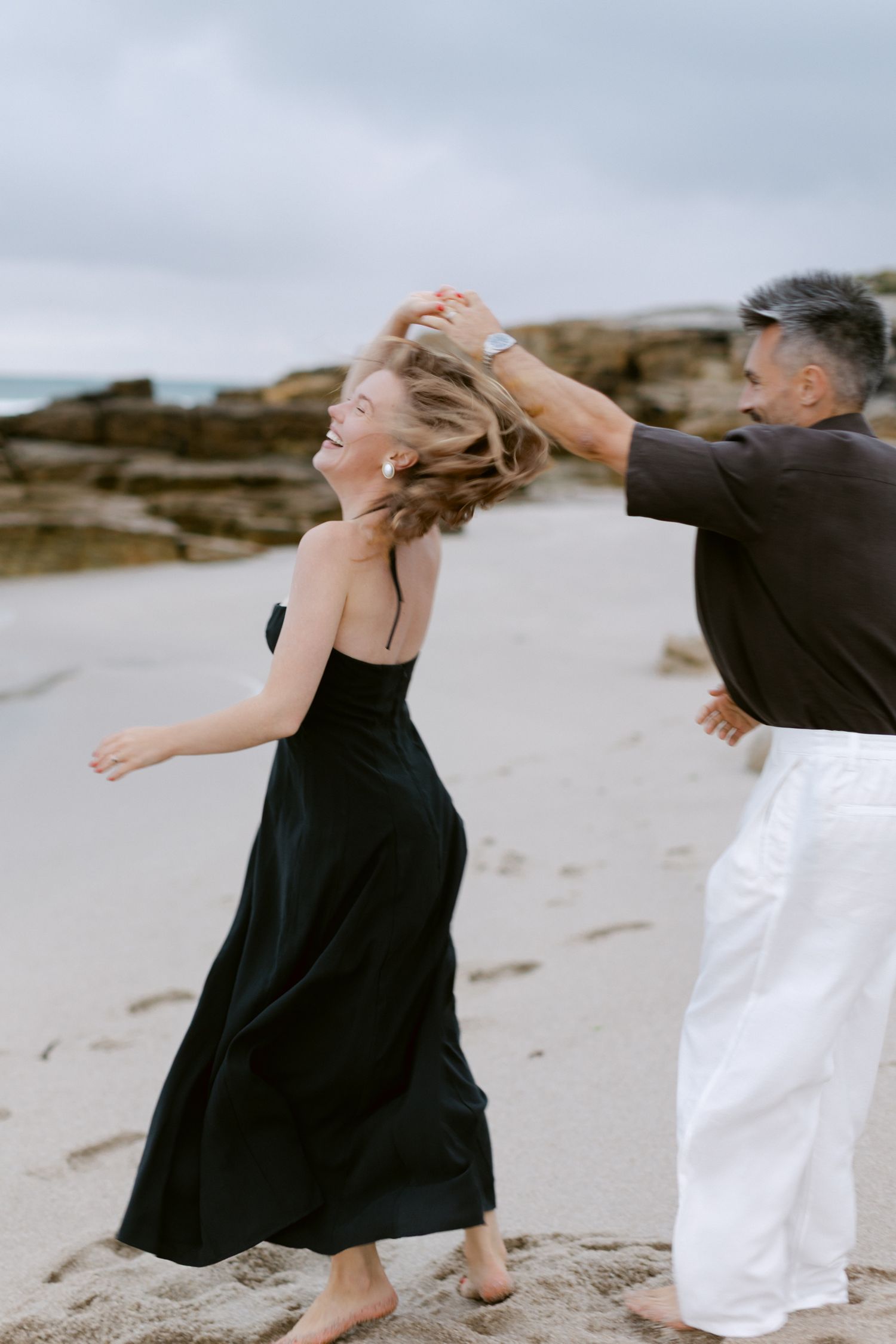 Couple dansant joyeusement sur une plage de sable, femme en robe longue noire se faisant tourner par un homme en pantalon blanc.