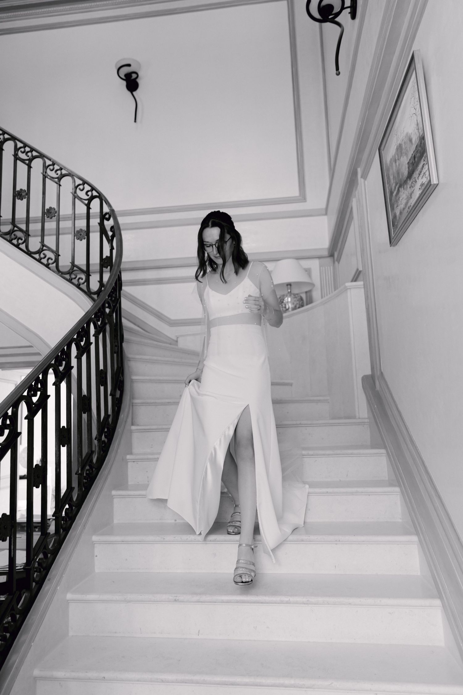 La mariée en robe blanche avec fente descend un escalier élégant avec une balustrade en fer forgé en photo noire et blanche.