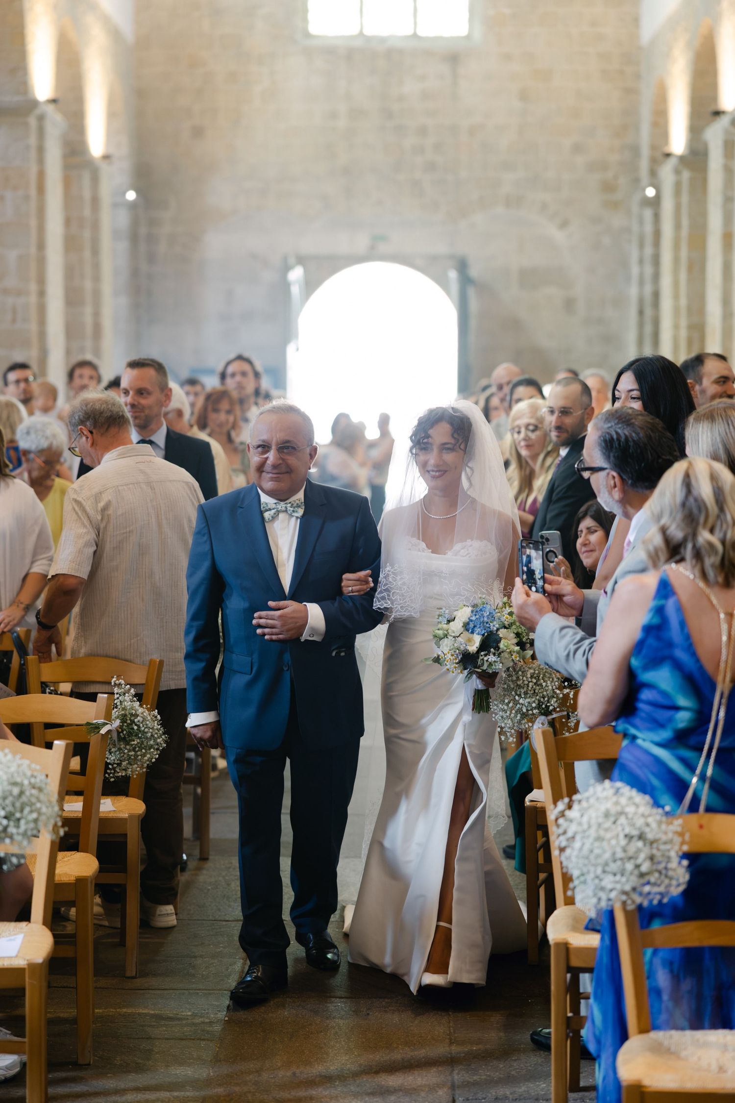 Bride walks down church aisle with older man in blue suit, guests seated in wooden chairs, stone walls, holding white bouquet.