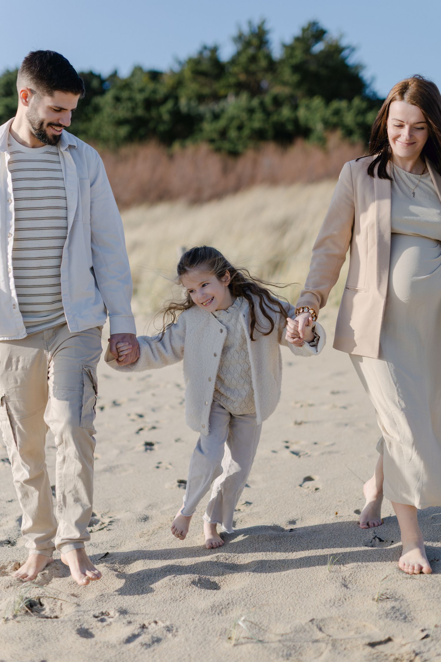 Famille joyeuse se tenant la main marchant pieds nus sur la plage de sable avec une mère enceinte et sa jeune fille.