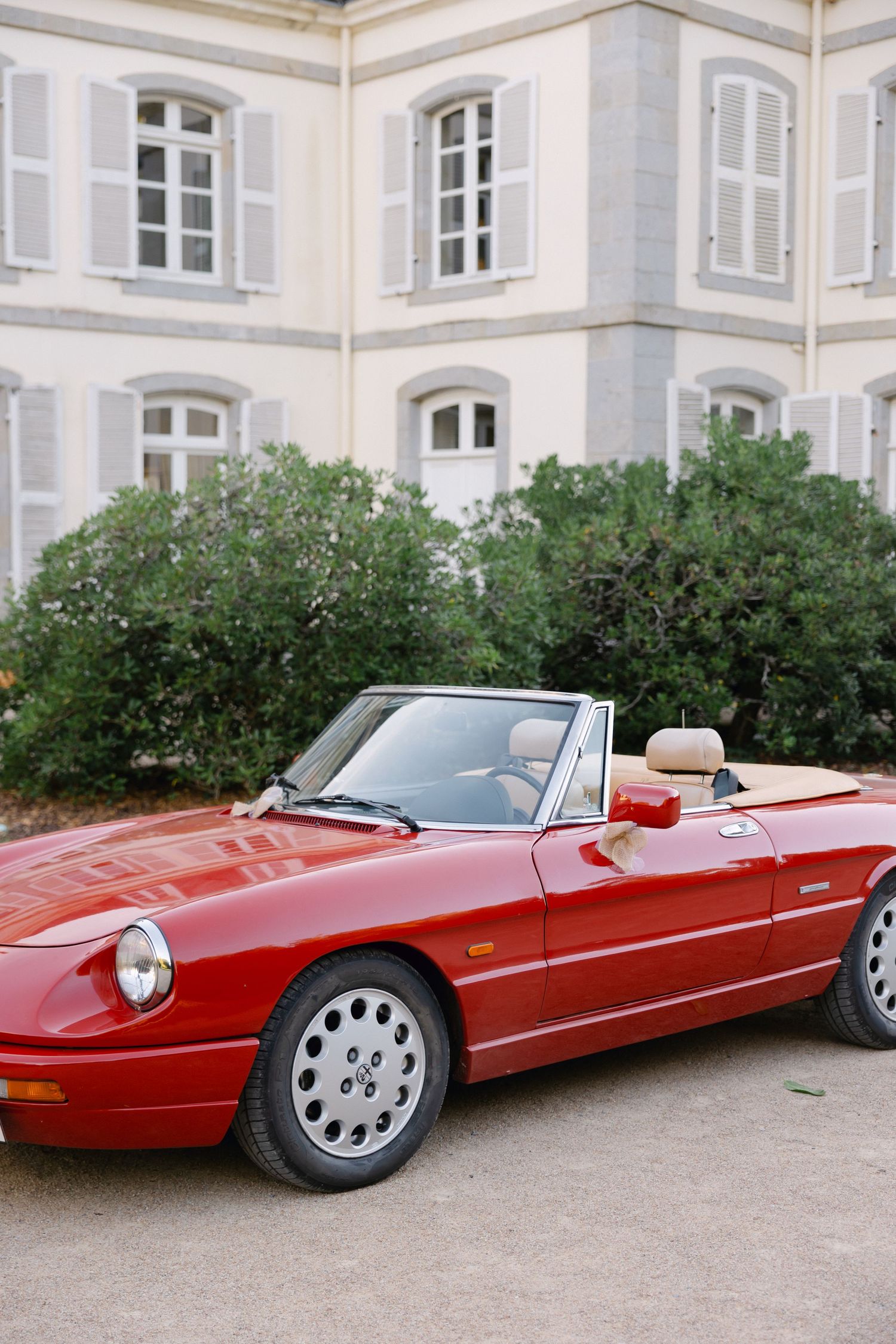 Cabriolet Alfa Romeo Spider rouge garée devant un bâtiment en pierre français classique avec des haies vertes luxuriantes.