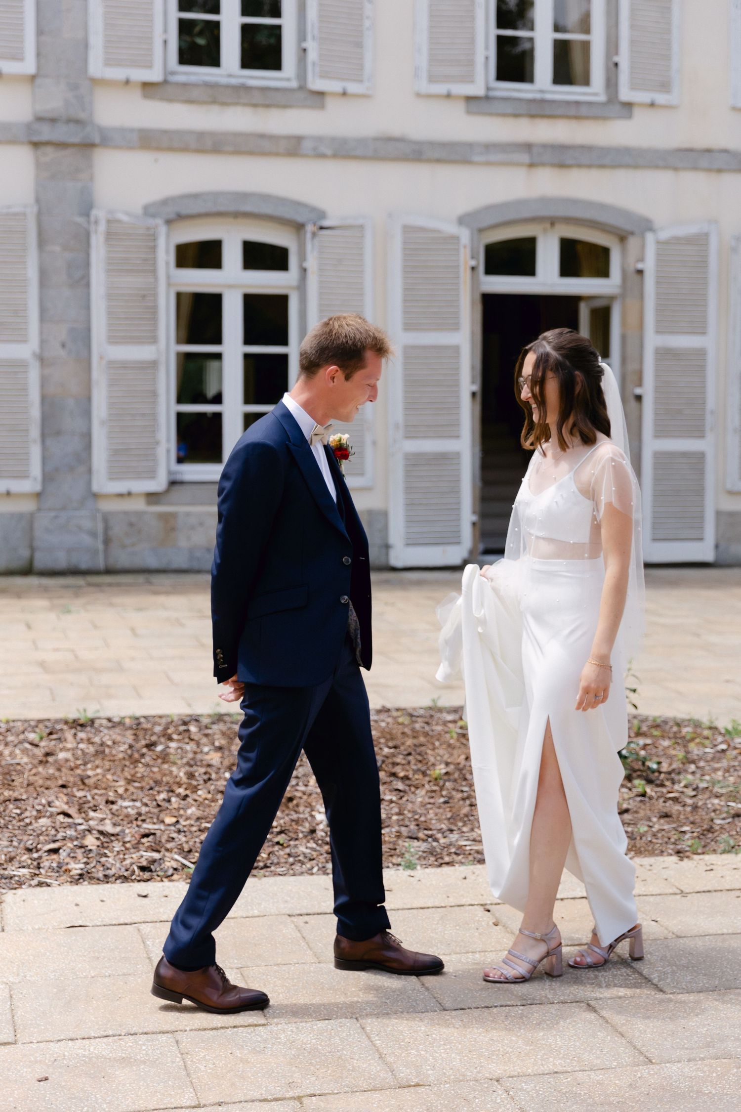 Une mariée en robe blanche et un marié en costume bleu marine marchent ensemble devant un élégant château français le jour de leur mariage.