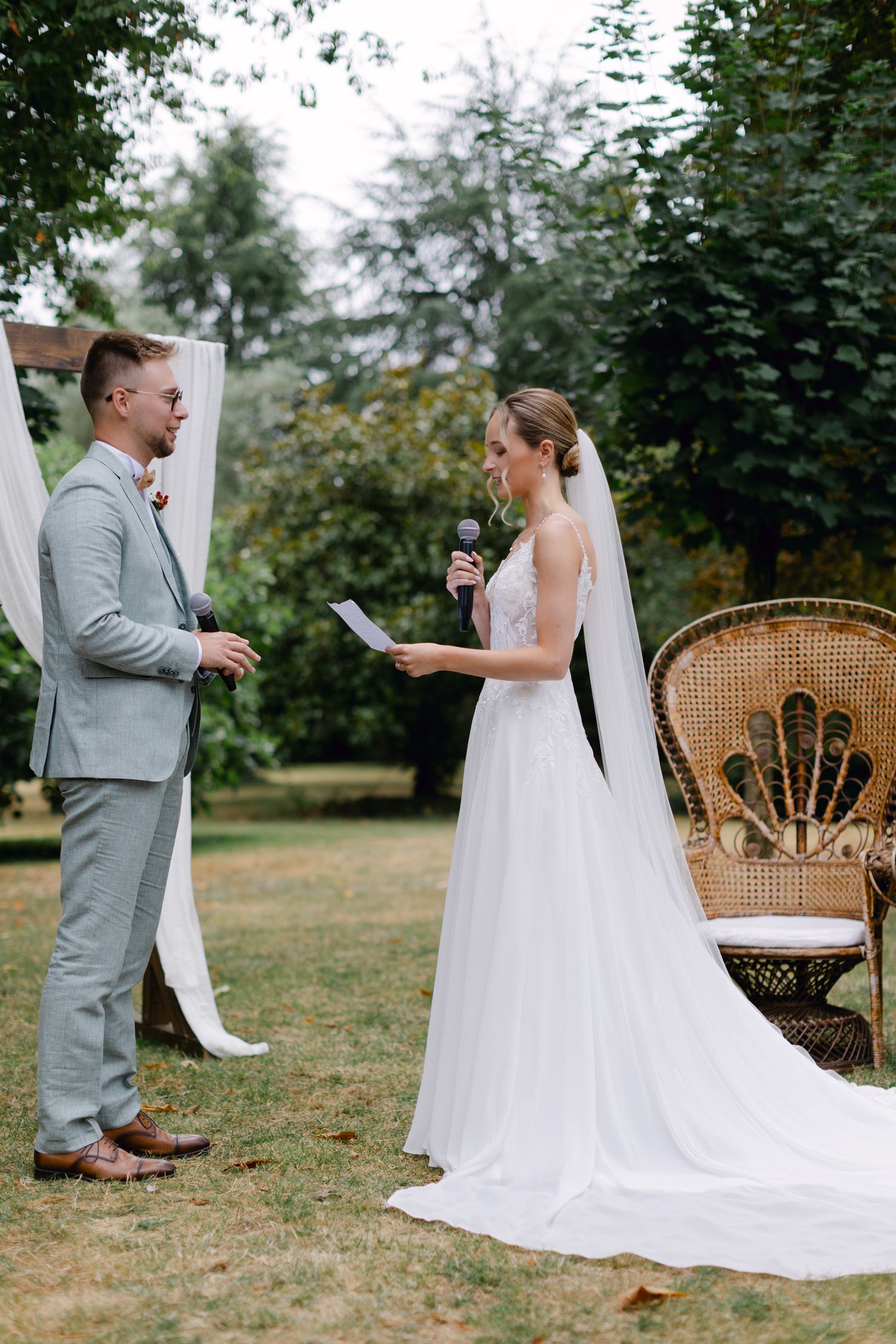 La mariée lit les vœux dans le microphone pendant que le marié écoute lors d'une cérémonie de mariage dans un jardin en plein air.