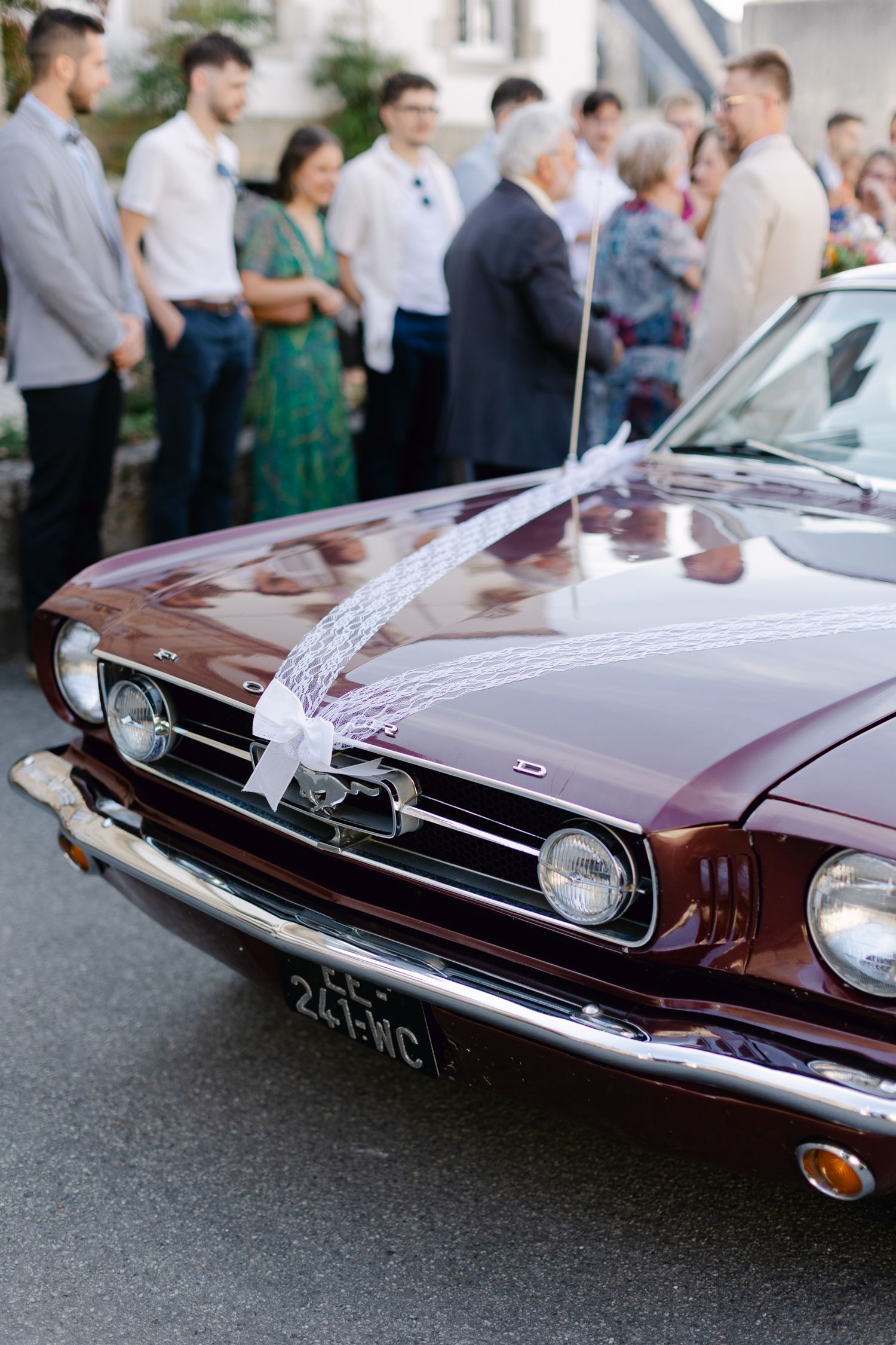 Ford Mustang bordeaux classique décorée d'un ruban de dentelle blanche garée à l'extérieur d'un lieu de mariage français.