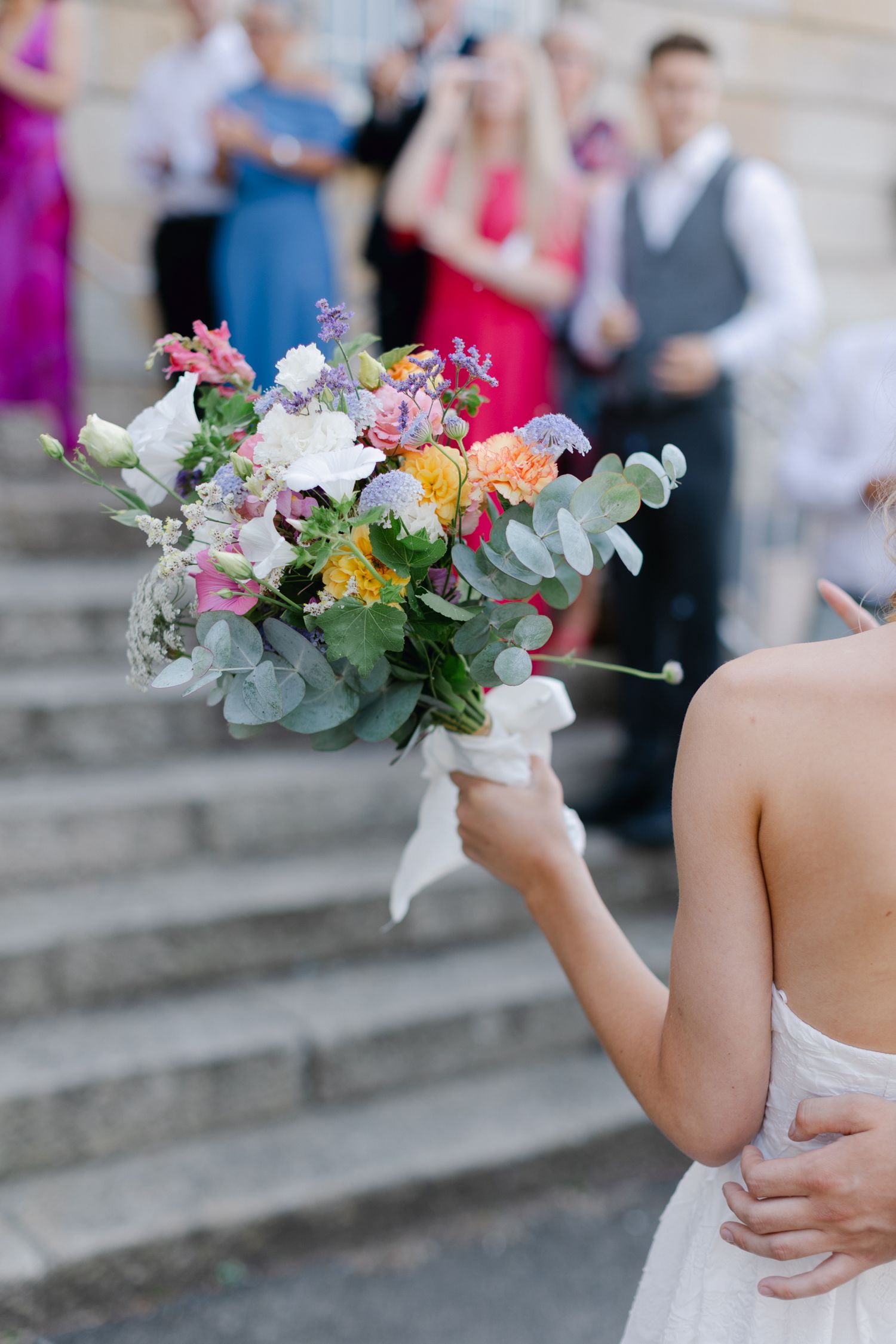 La mariée tient un bouquet de mariage vibrant de fleurs mélangées et d'eucalyptus tandis que les invités se rassemblent sur des marches.