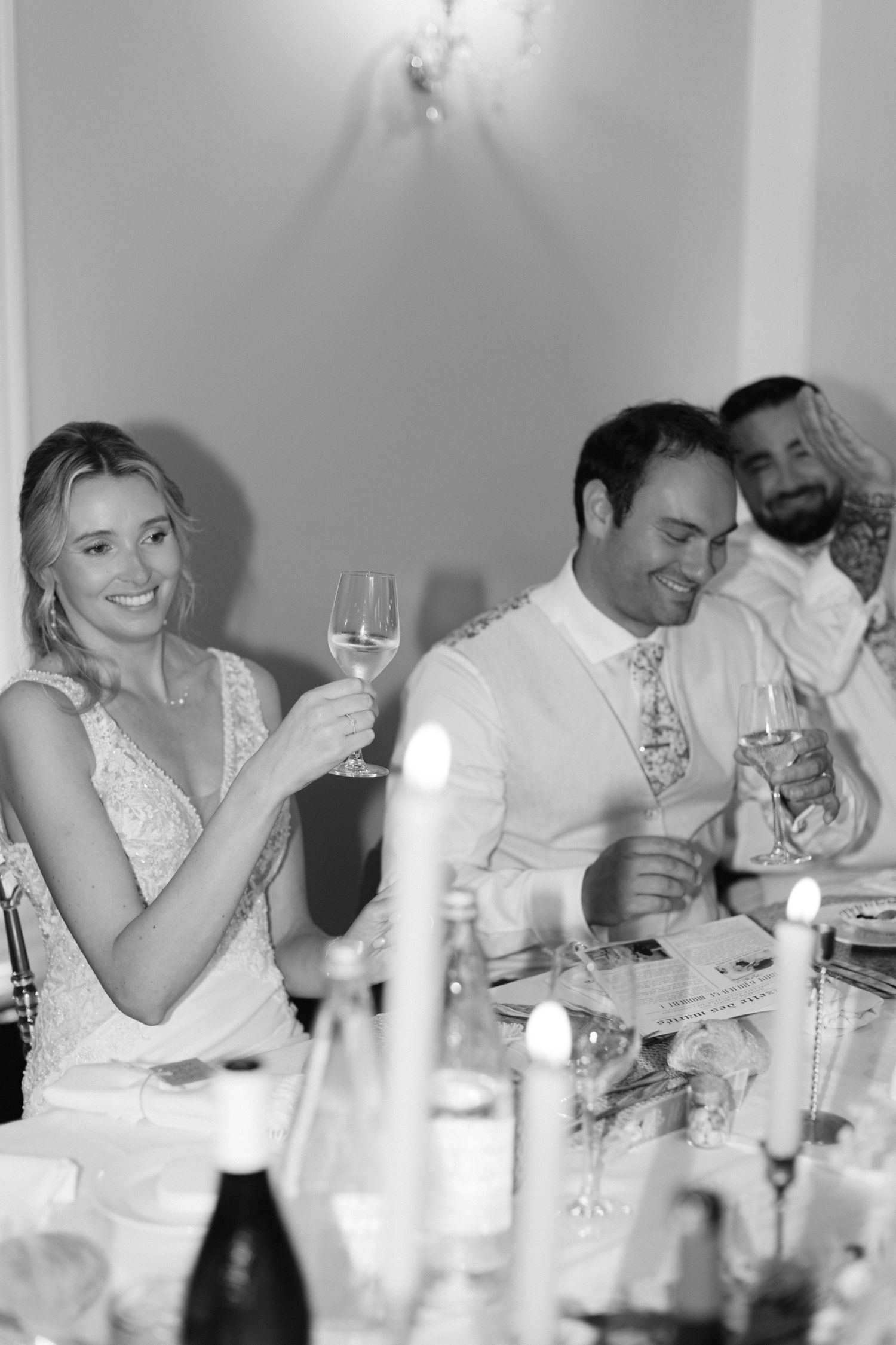 Photo en noir et blanc d'une mariée souriante levant un verre de champagne lors d'un dîner de réception de mariage.